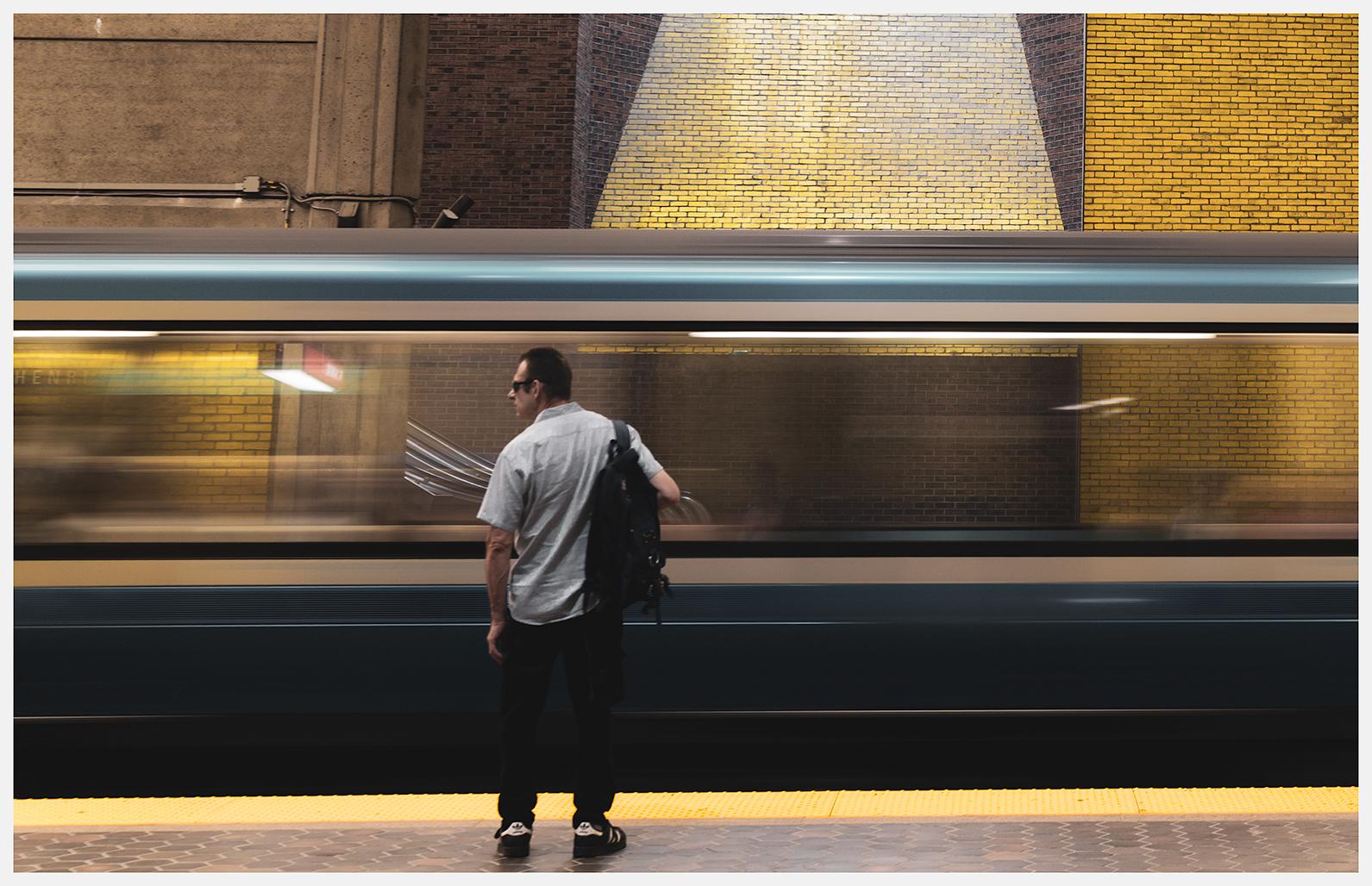 An image of a metro station (St-Henri in Montreal) with a man in the foreground wearing dark glasses, a light, short-sleeved shirt, dark pants and sneakers and carrying a black backpack. He's standing in front of a yellow safety line and looking to the left toward something outside of the frame as a blurred blue and white-trimmed train passes him by. The windows of the train blur together, seeming to form one long window through which the yellow and red bricks, concrete walls, and the escalator of the other side side of the station can clearly be seen.