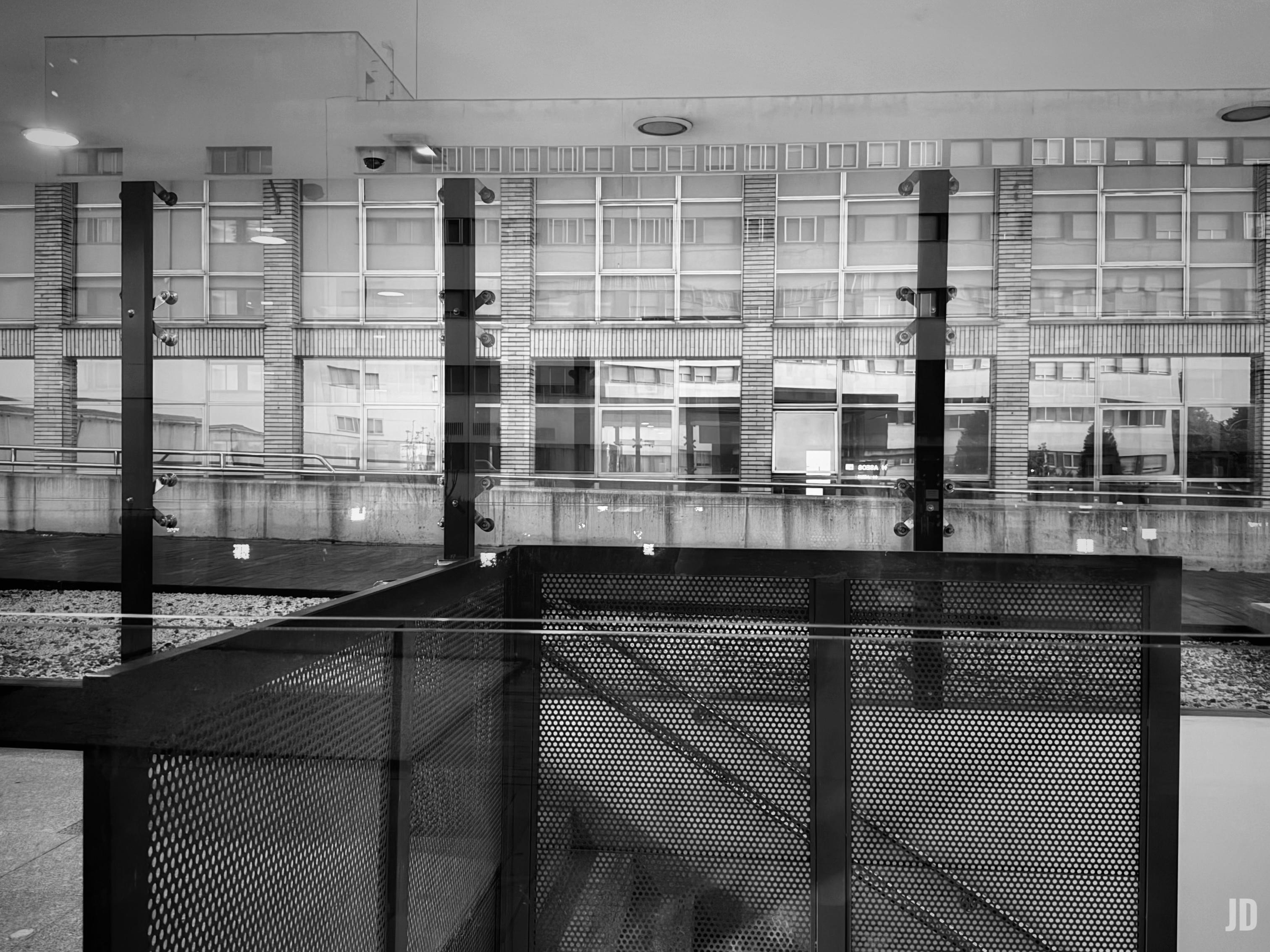 La fotografía en blanco y negro muestra una perspectiva arquitectónica moderna que combina capas visuales y reflejos.
En primer plano destaca una barandilla de paneles metálicos perforados junto a unas escaleras que descienden. Detrás de esta estructura, se observa un gran ventanal de vidrio con soportes metálicos verticales, sobre el cual se reflejan las ventanas cuadradas y la fachada de un edificio contemporáneo, creando una superposición geométrica entre el interior y el exterior.