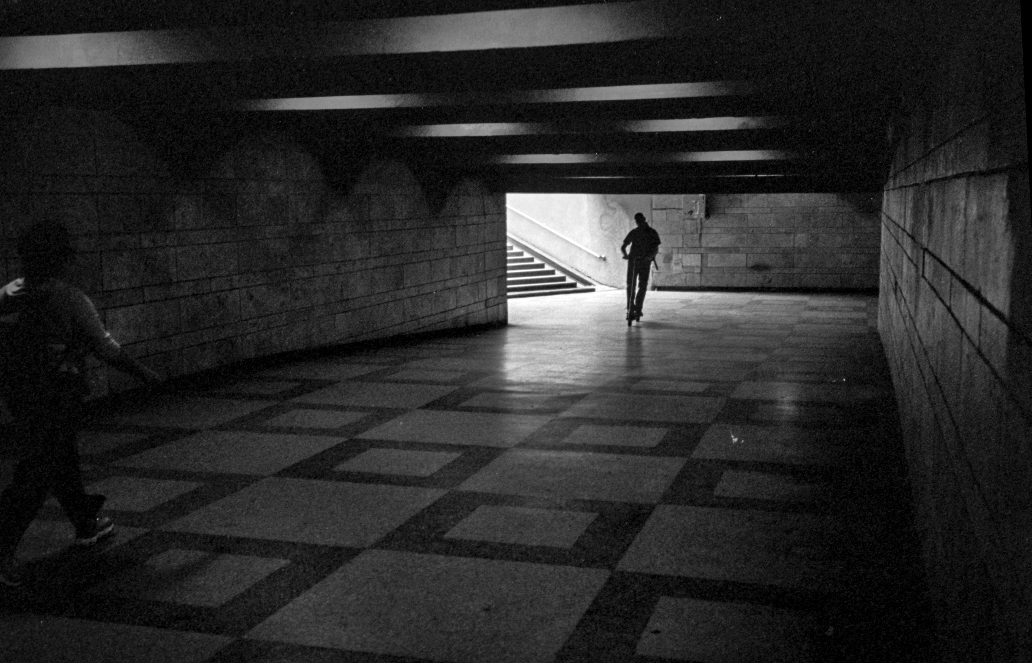 A young man rides an electric scooter through an underground pedestrian passage in central Lviv, Ukraine. The dimly lit passage has tiled walls and a geometric tiled floor, with bright daylight visible at the exit ahead.