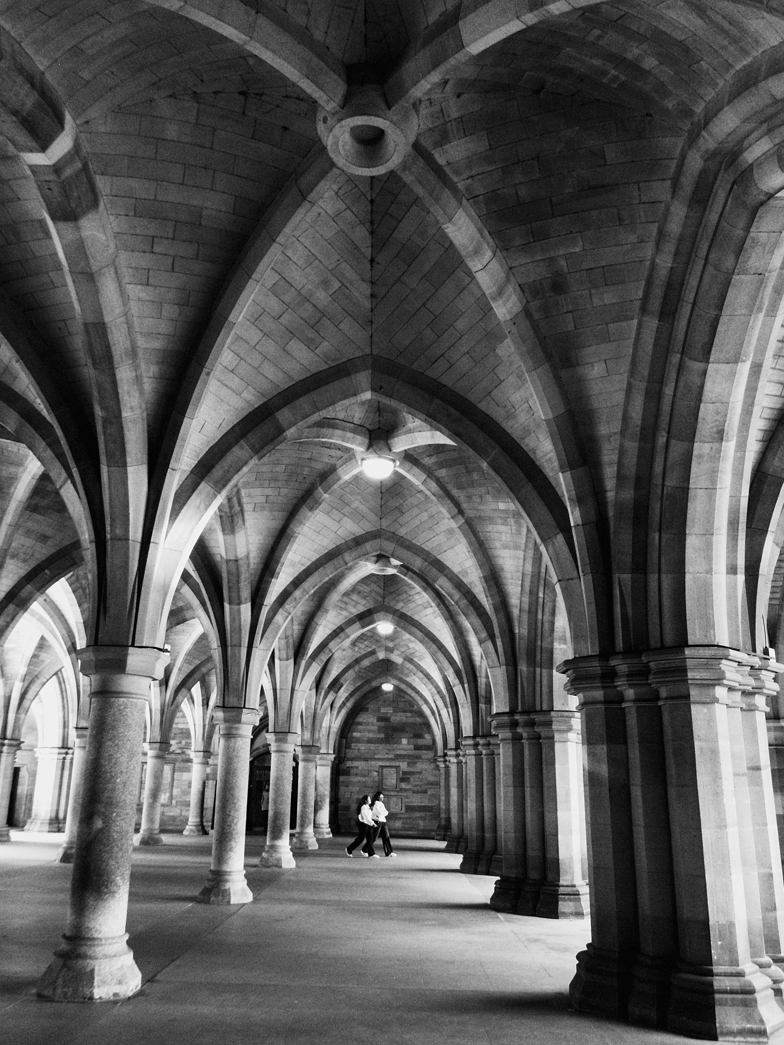 Two people walk through the beautiful stone cloisters at Glasgow University, where the ancient stonework rises in soaring archways to a geometrically designed, curved ceiling