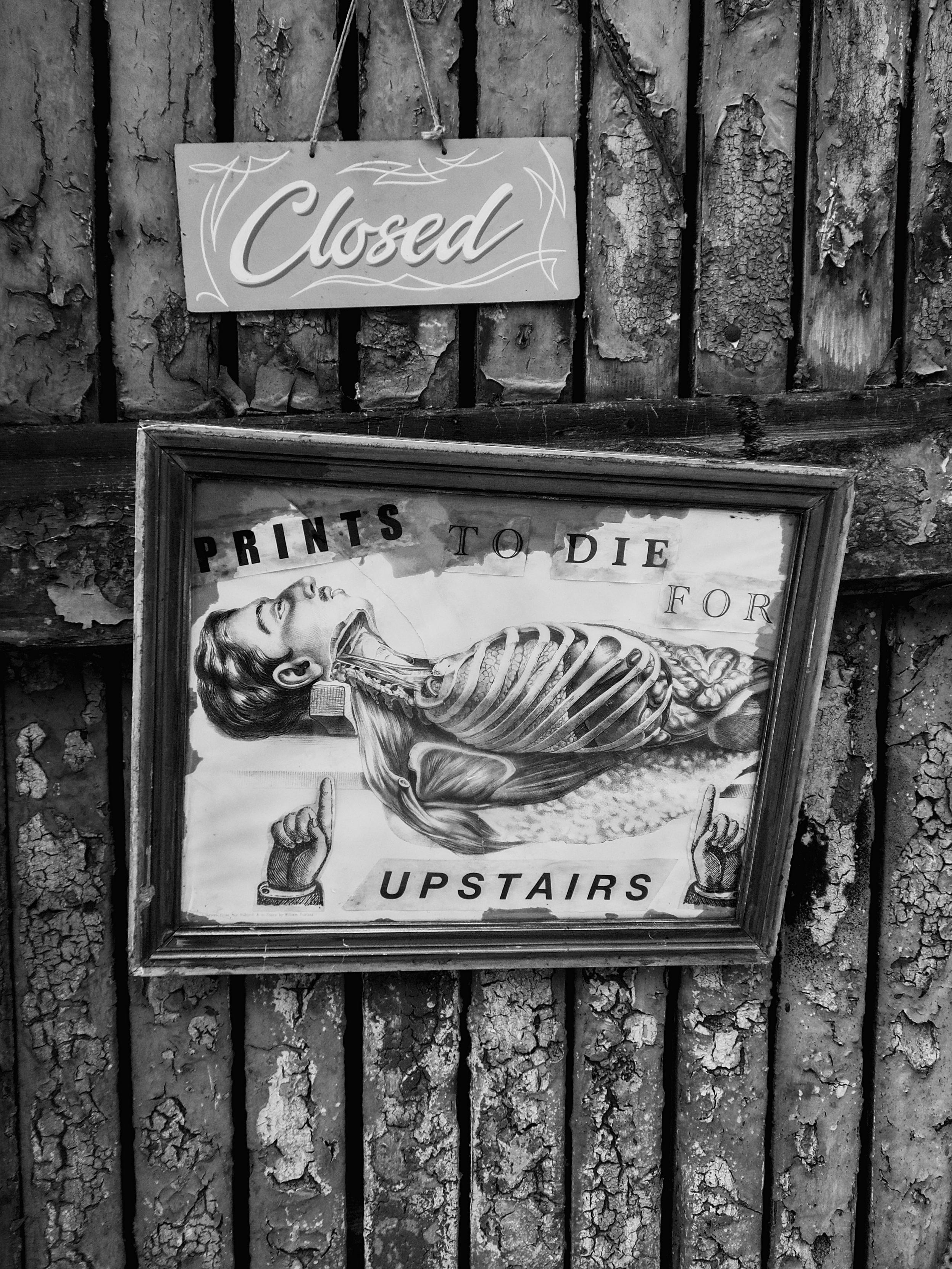 Two signs, one reading 'Closed' in freehand-style script and the other reading 'Prints to die for, upstairs', hang slightly askew on an old wooden door covered in cracked and peeling paint in a lane full of small independent businesses in the Finnieston area of Glasgow