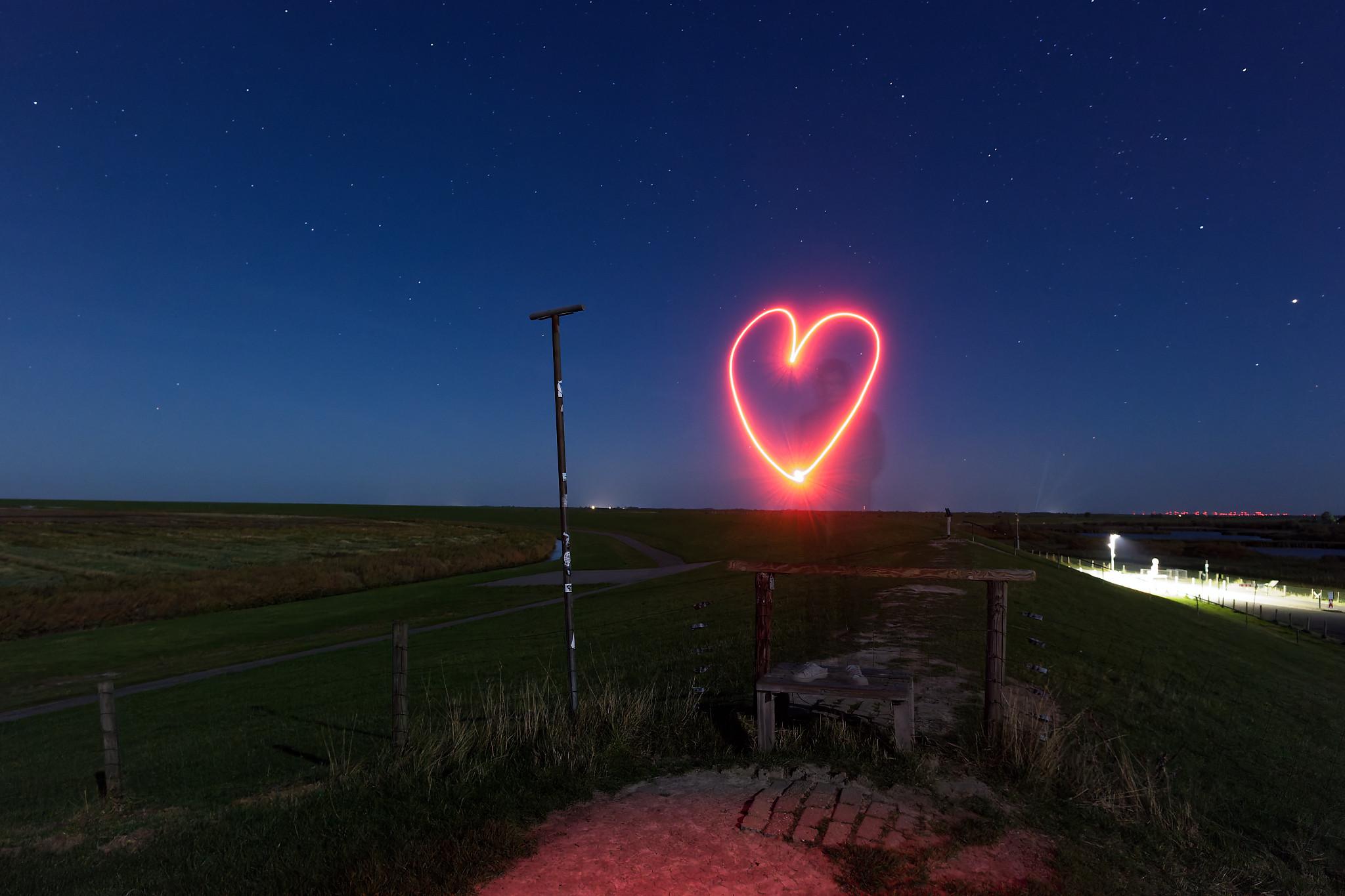 Nachtaufnahme auf einem Deich in Ostfriesland. Im Hintergrund ist der Himmel mit Sternen sichtbar. Im Vordergrund malt jemand mit einer roten LED-Taschenlampe ein Herz aus Licht. Die Person selber ist dabei durch die Langzeitbelichtung fast unsichtbar.