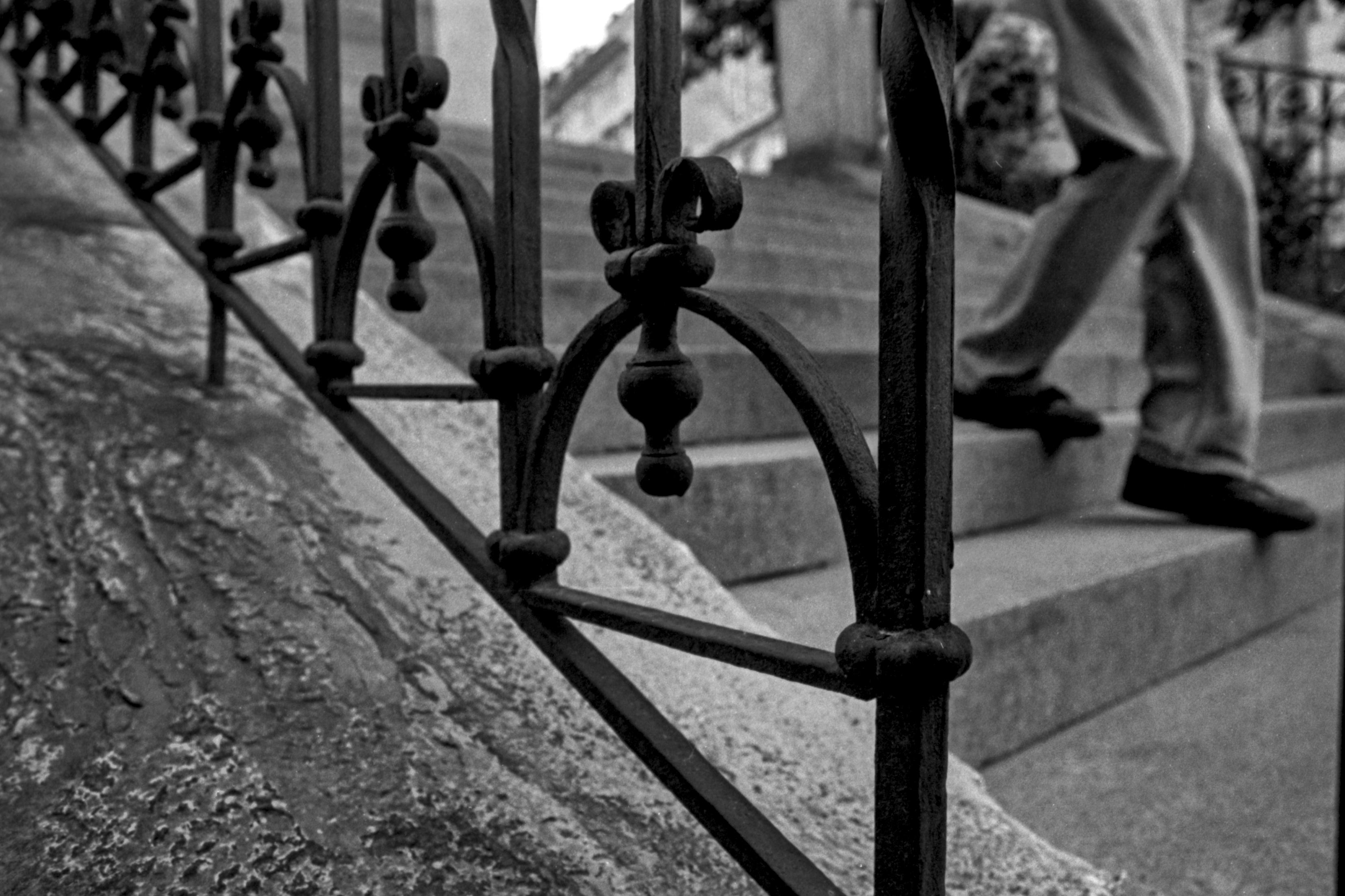 A black-and-white photograph of an ornate wrought-iron railing alongside a stone staircase, with a person descending the steps in the background. The shallow depth of field and strong textures create a moody, vintage atmosphere.