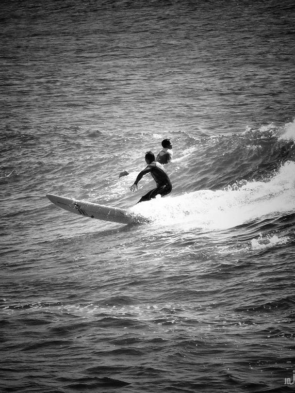 La imagen en blanco y negro muestra a dos personas surfeando una ola en el mar. La persona en primer plano está de pie sobre la tabla, inclinada hacia adelante mientras surfea, mientras que la otra está un poco más atrás, también sobre la ola. El agua forma espuma blanca alrededor de la tabla que va en movimiento.