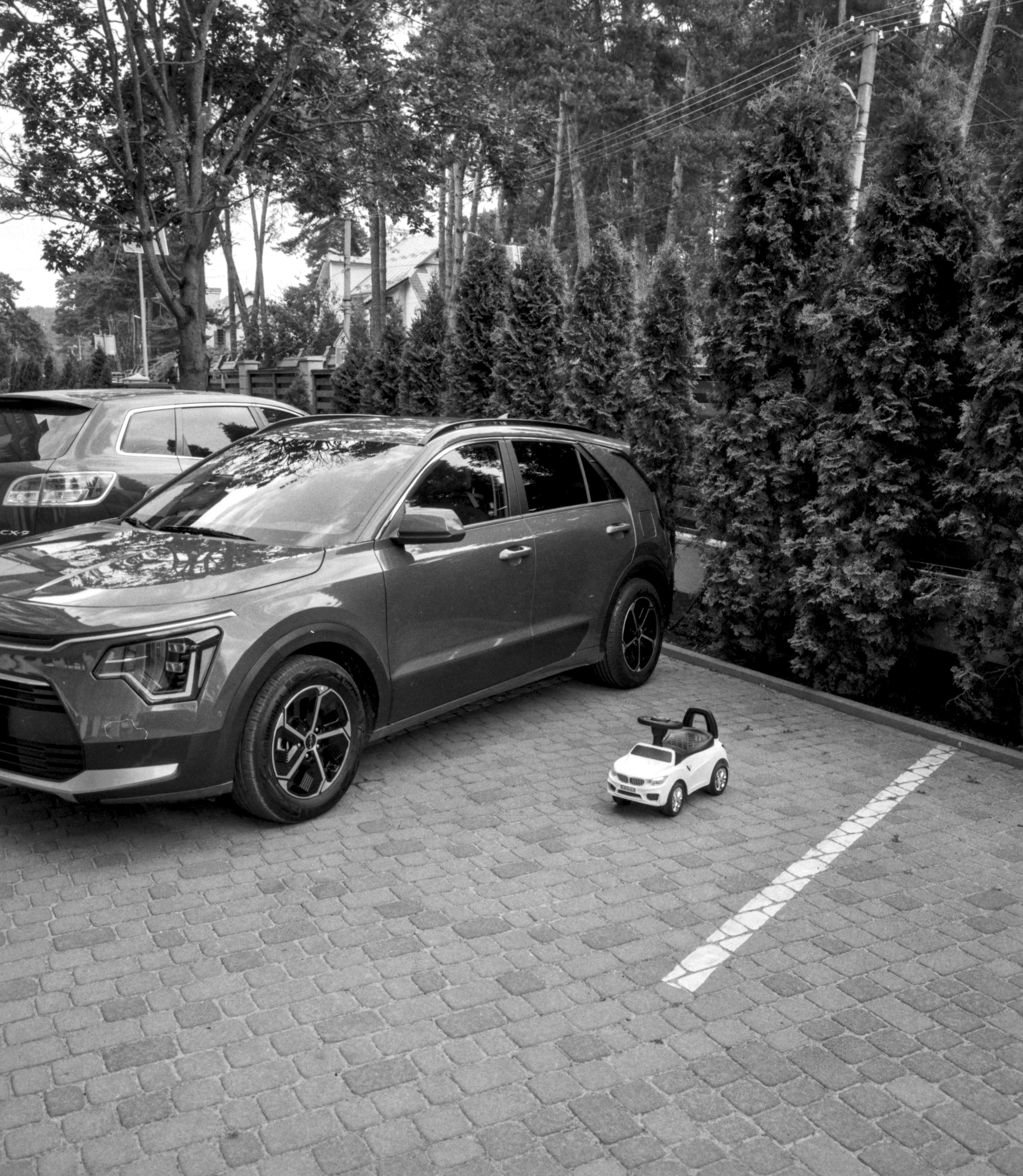 A black-and-white photograph of a small toy car parked beside a full-size SUV in a paved parking area, creating a playful visual contrast. Tall evergreen trees, utility poles, overhead wires, and residential houses form the background.