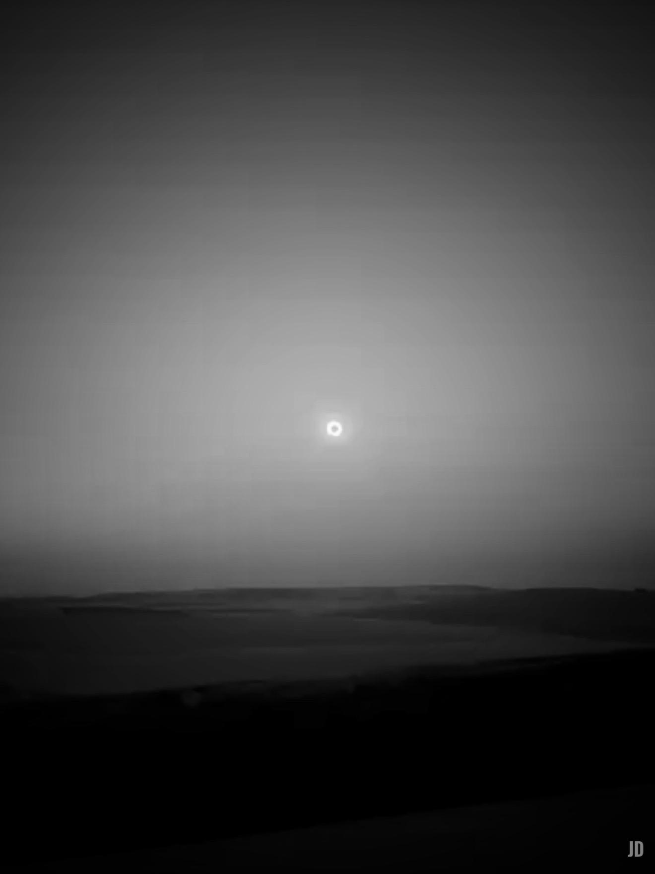 La imagen muestra un paisaje en blanco y negro con un horizonte llano y oscuro en la parte inferior, posiblemente campos o colinas bajas. En el cielo, destaca un sol cubierto por la luna totalmente, creando un efecto de halo brillante en el centro de la escena. La atmósfera es serena y ligeramente misteriosa.