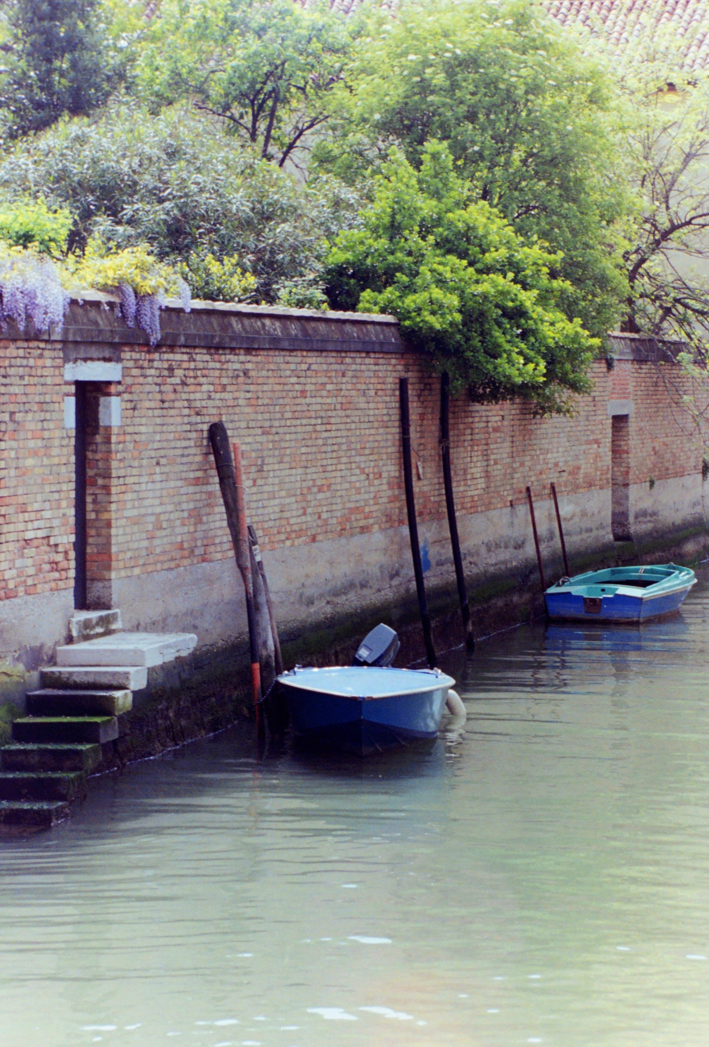 A photograph taken with film shows a tranquil canal in Venice, where two boats are moored. The canal is bordered on one side by a long exposed brick wall with two gates, the nearest of which has a built-in staircase leading down to the water. Beyond the wall, a large garden is visible, above which rise the tops of numerous trees.