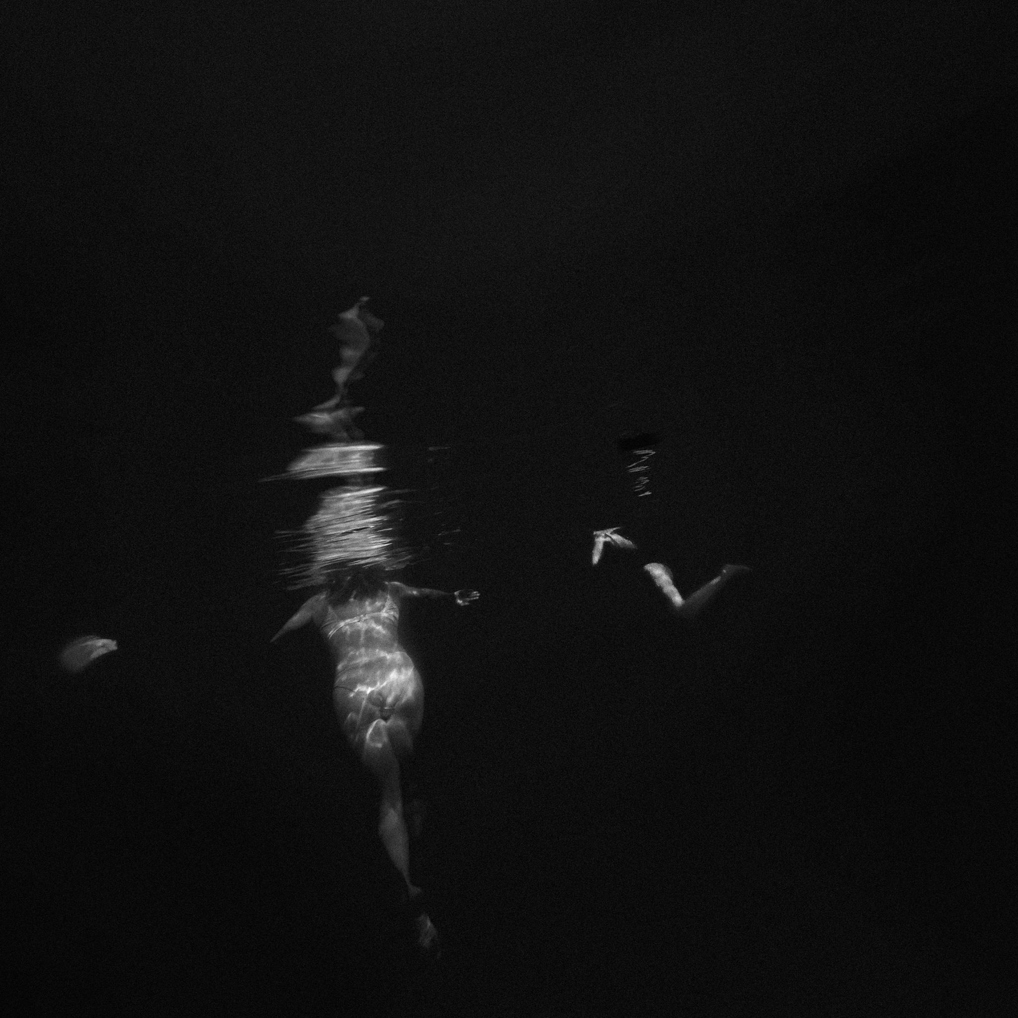 Black and white underwater photograph of a human figure partially illuminated beneath dark water, with reflections and distortions breaking up the body and surrounding forms.