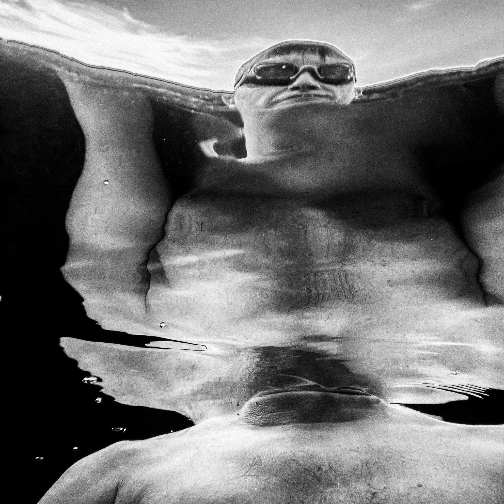 Black and white underwater photograph of a person wearing sunglasses at the waterline, with the submerged body heavily distorted and duplicated by ripples and reflections.