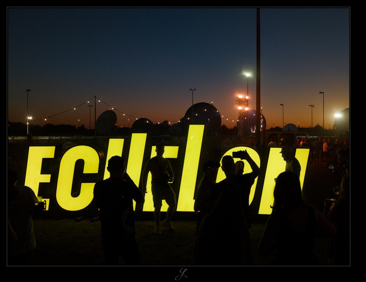 A yellow neon sign reading ECHELON in all capital letters. People are posing in front of the neon sign to take a good photo. The people posing in front of the sign and those taking photos with their cell phones are visible as dark silhouettes against the bright yellow lettering. Behind them, radar domes and lampposts rise as black silhouettes against the dark orange to dark-blue evening sky.

AI disclaimer: Using my work, its meta data, written or derived description to create media with or train AI based systems is prohibited.