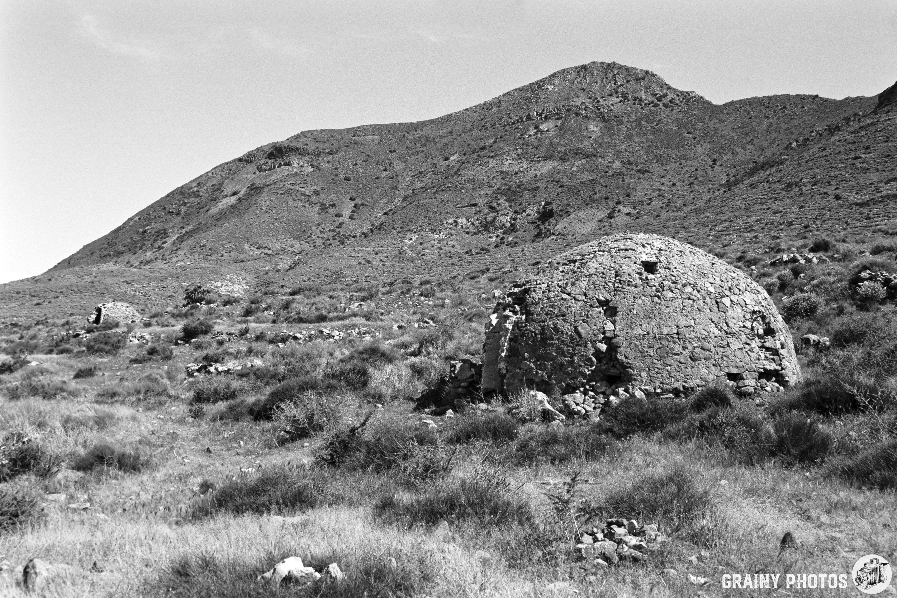 A black and white landscape featuring rounded stone structures amidst grassy terrain, set against a backdrop of a mountain. The scene conveys a sense of history and solitude in a natural environment.