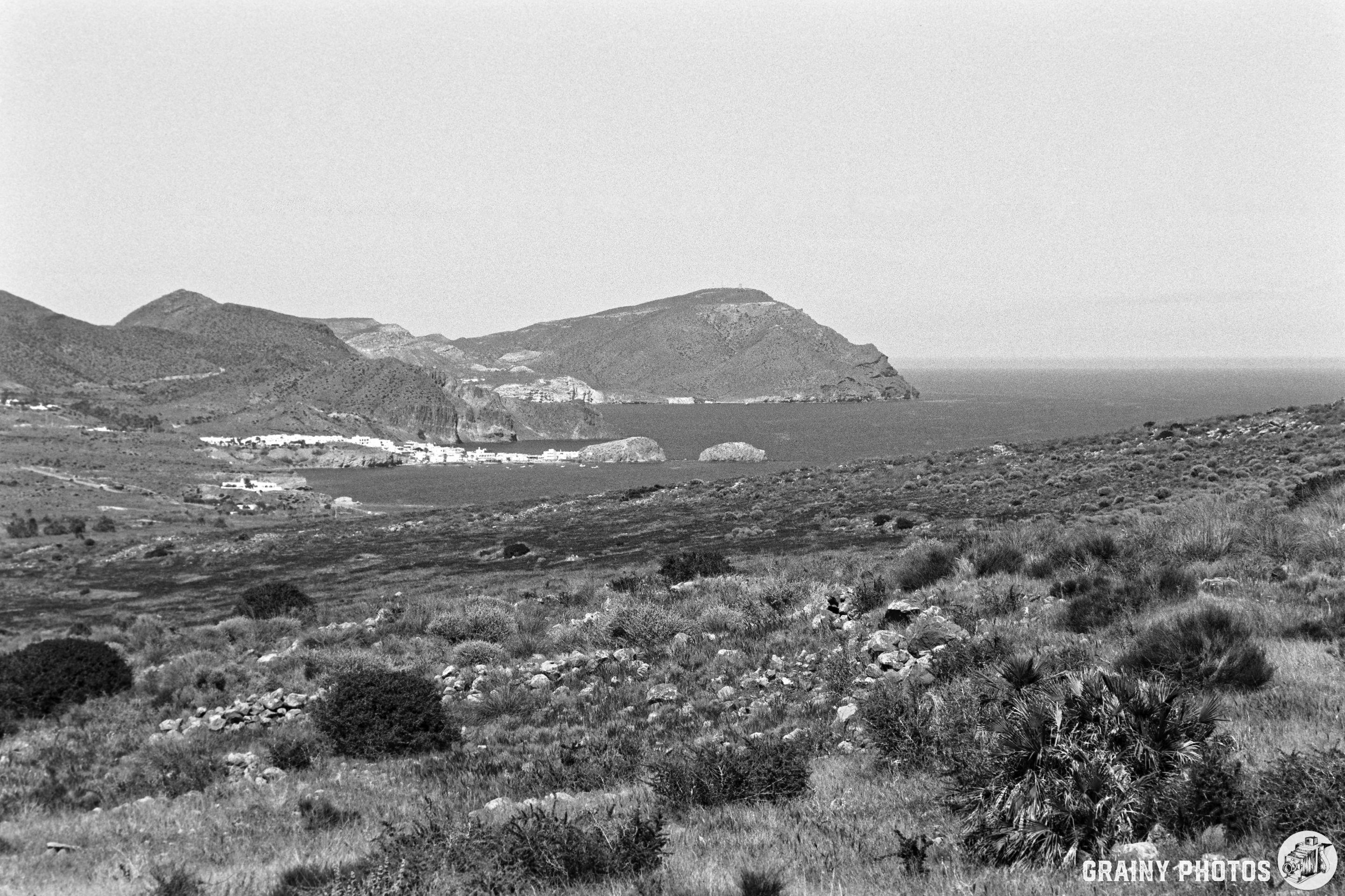 A serene coastal landscape features rolling hills and rocky formations, overlooking a calm sea. The scene is captured in black and white, highlighting the natural contours and textures of the terrain.