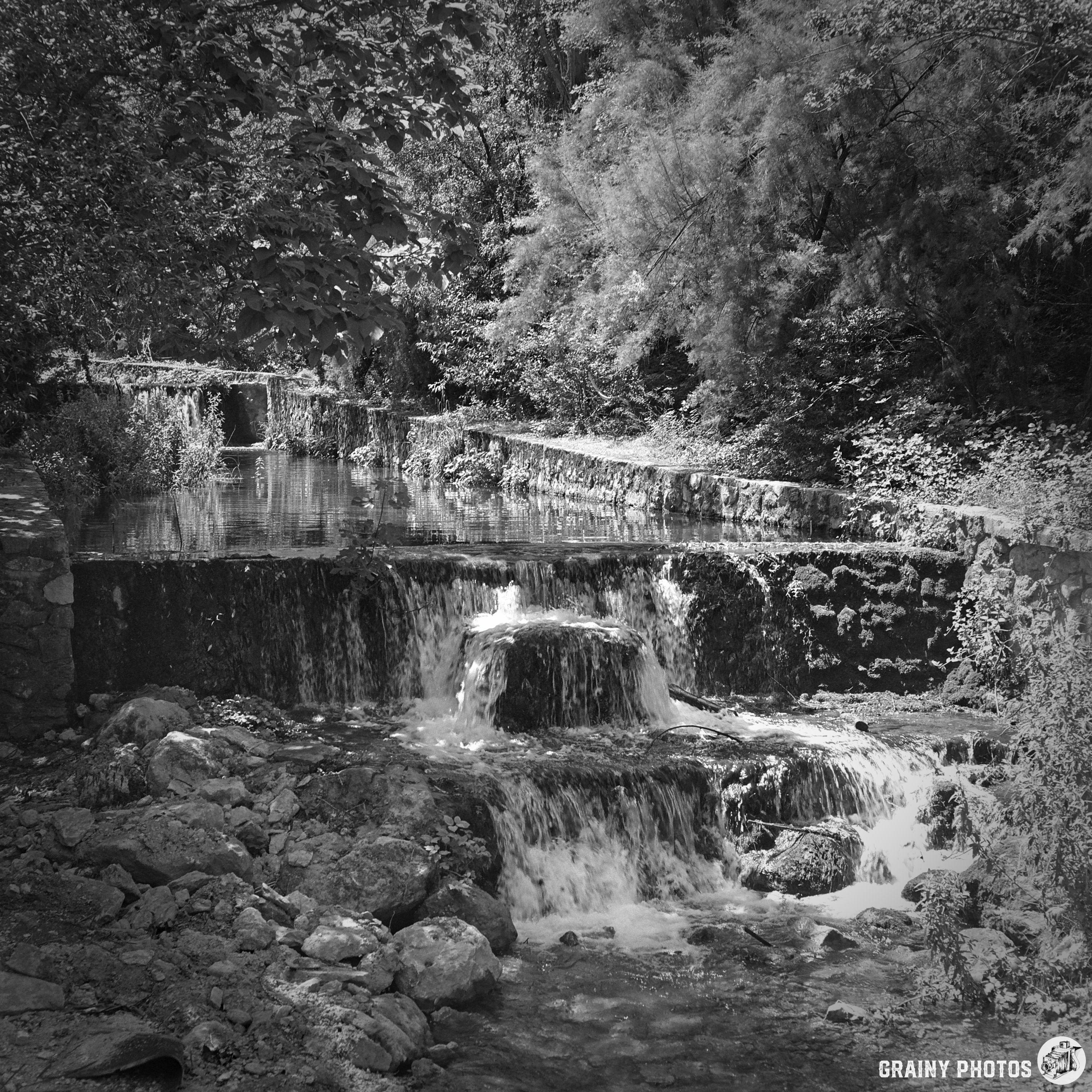 A serene black and white image of cascading water over rocks, surrounded by lush greenery and trees, creating a tranquil natural scene. The calm flow of the water adds to the peaceful atmosphere of the landscape.