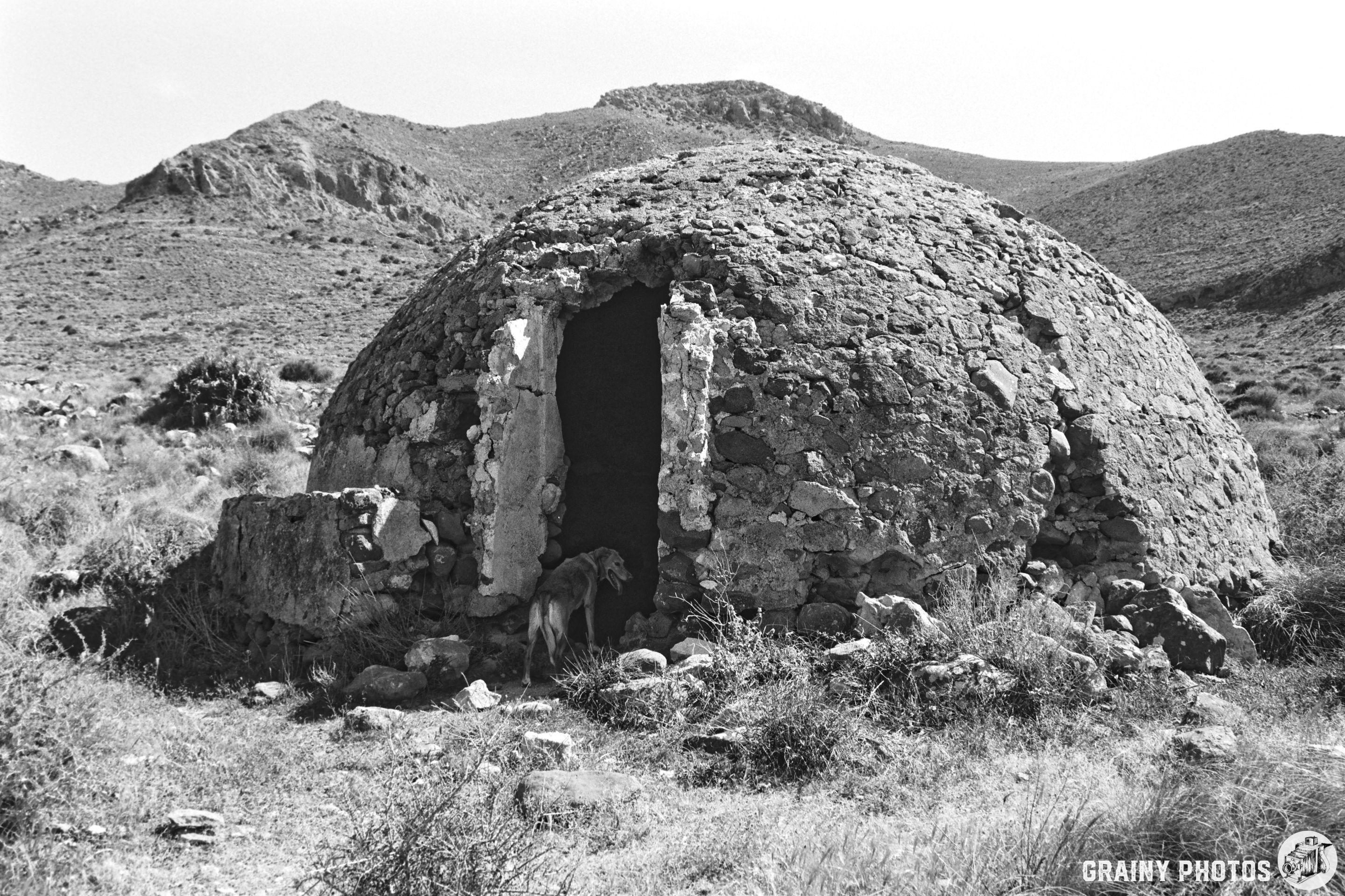 A weathered stone structure resembling an igloo stands in a dry, mountainous landscape. A dog is seen near the entrance, with sparse vegetation surrounding the area, all captured in black and white.