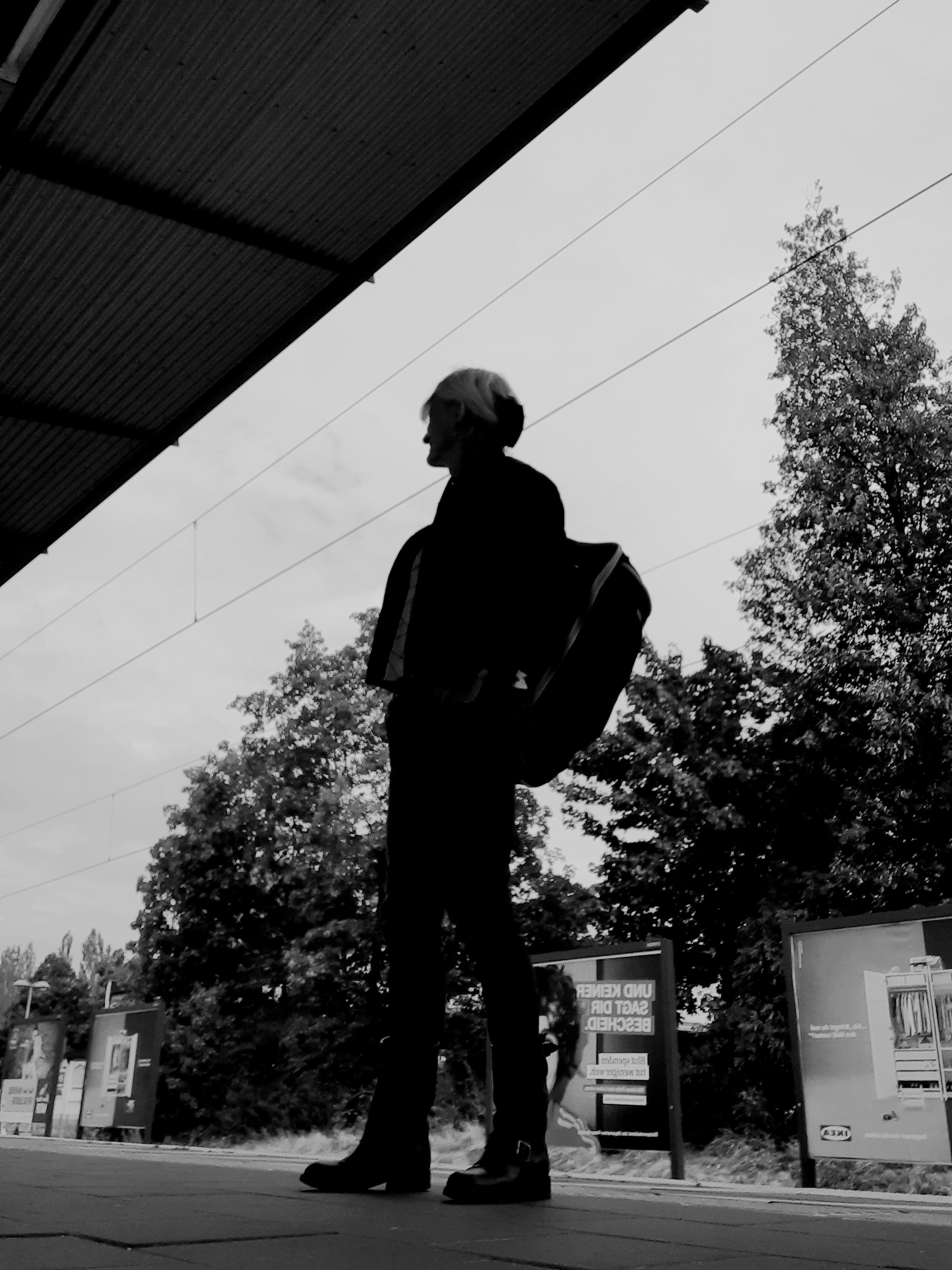 The outline of a person standing on a train platform. She wears heavy shoes and carries a backpack. In her back a row of trees and billboards