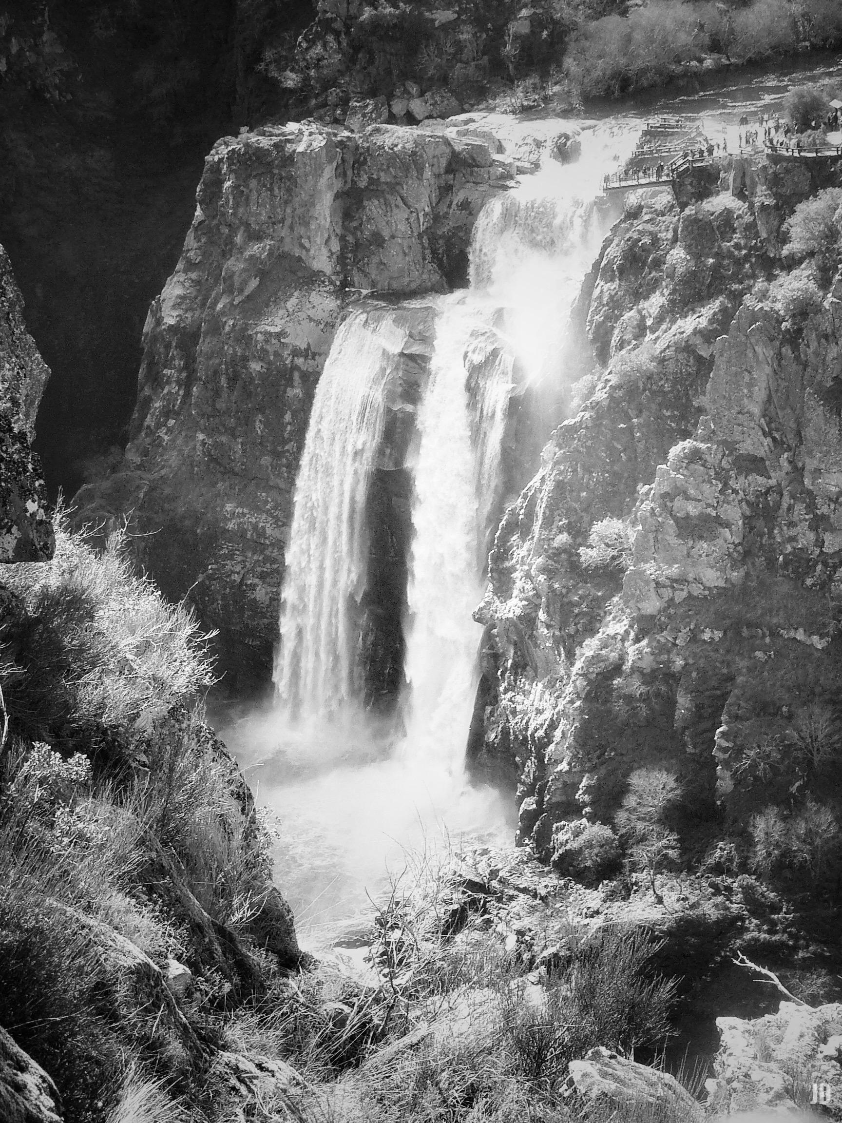 La imagen muestra una impresionante cascada cayendo entre rocas escarpadas. El agua se precipita en dos saltos principales, rodeada de vegetación silvestre y acantilados. En la parte superior derecha se observa una pasarela con varias personas contemplando el paisaje. La escena está en blanco y negro, lo que resalta los contrastes entre el agua, las rocas y la vegetación.
