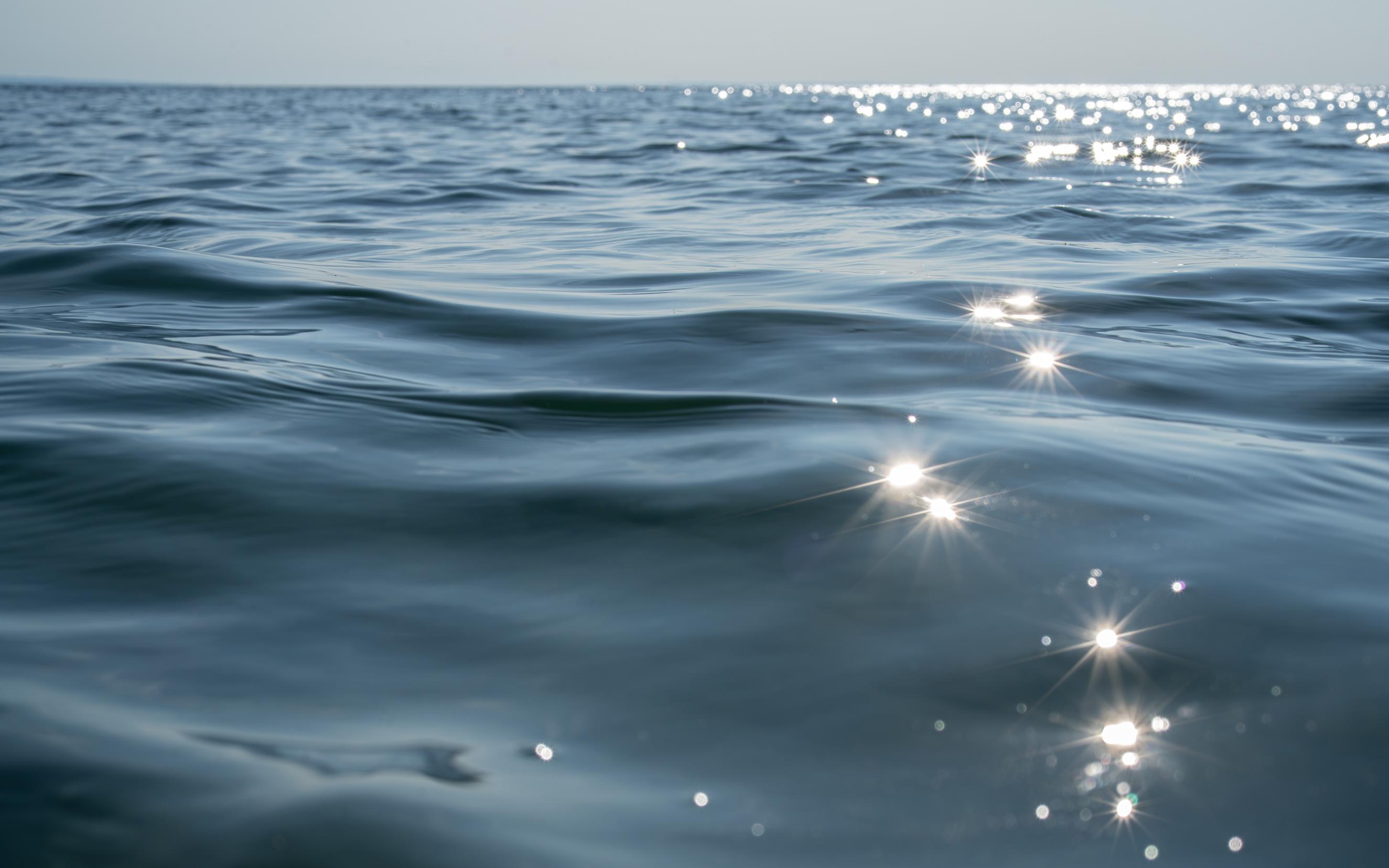 An close-up photo of a blue lake with gentle ripples taken in the water late in the afternoon. On the right side of the image, a line of sparkling sun star reflections. In the distance, more glittering reflections on the surface of the water. At the top of the image, a line of blue sky.