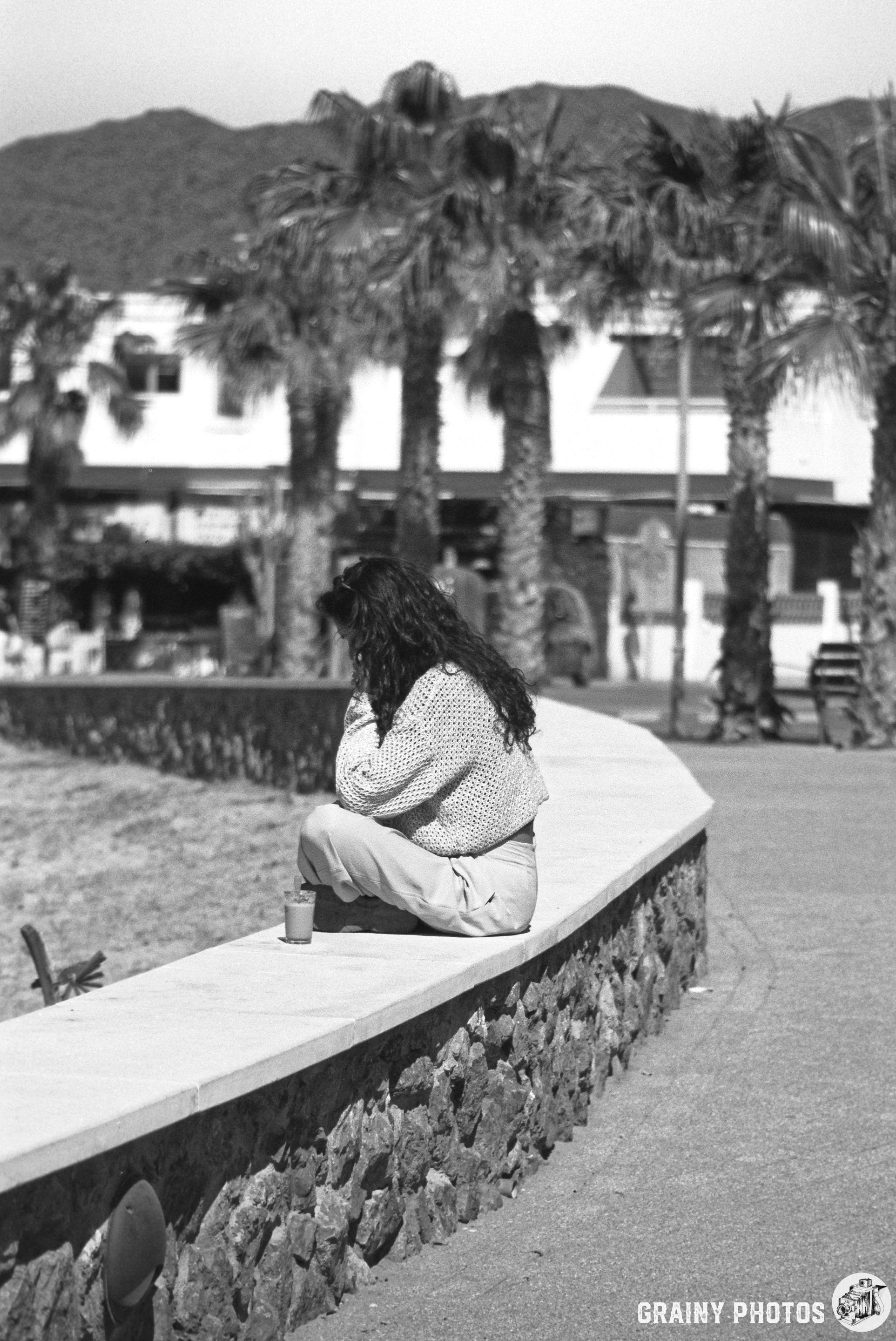 A woman with long hair sits on a stone wall, gazing thoughtfully into the distance. Palm trees and a buildings are visible in the background,. The image is captured in black and white.