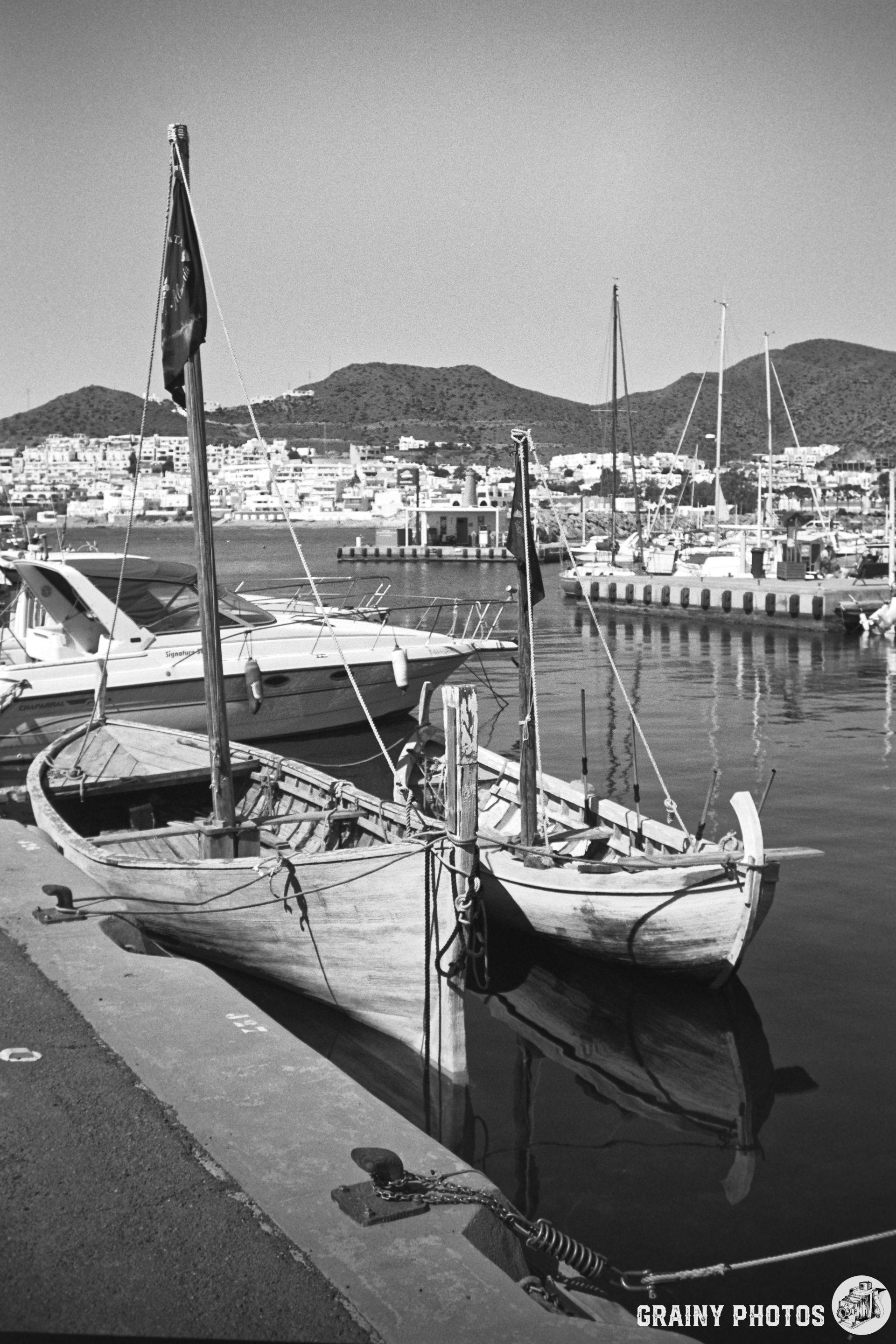A peaceful San José marina scene featuring two wooden boats docked at the pier, surrounded by a backdrop of distant hills and a bustling town. The image is in black and white, emphasizing the tranquil waters and the classic charm of the boats.
