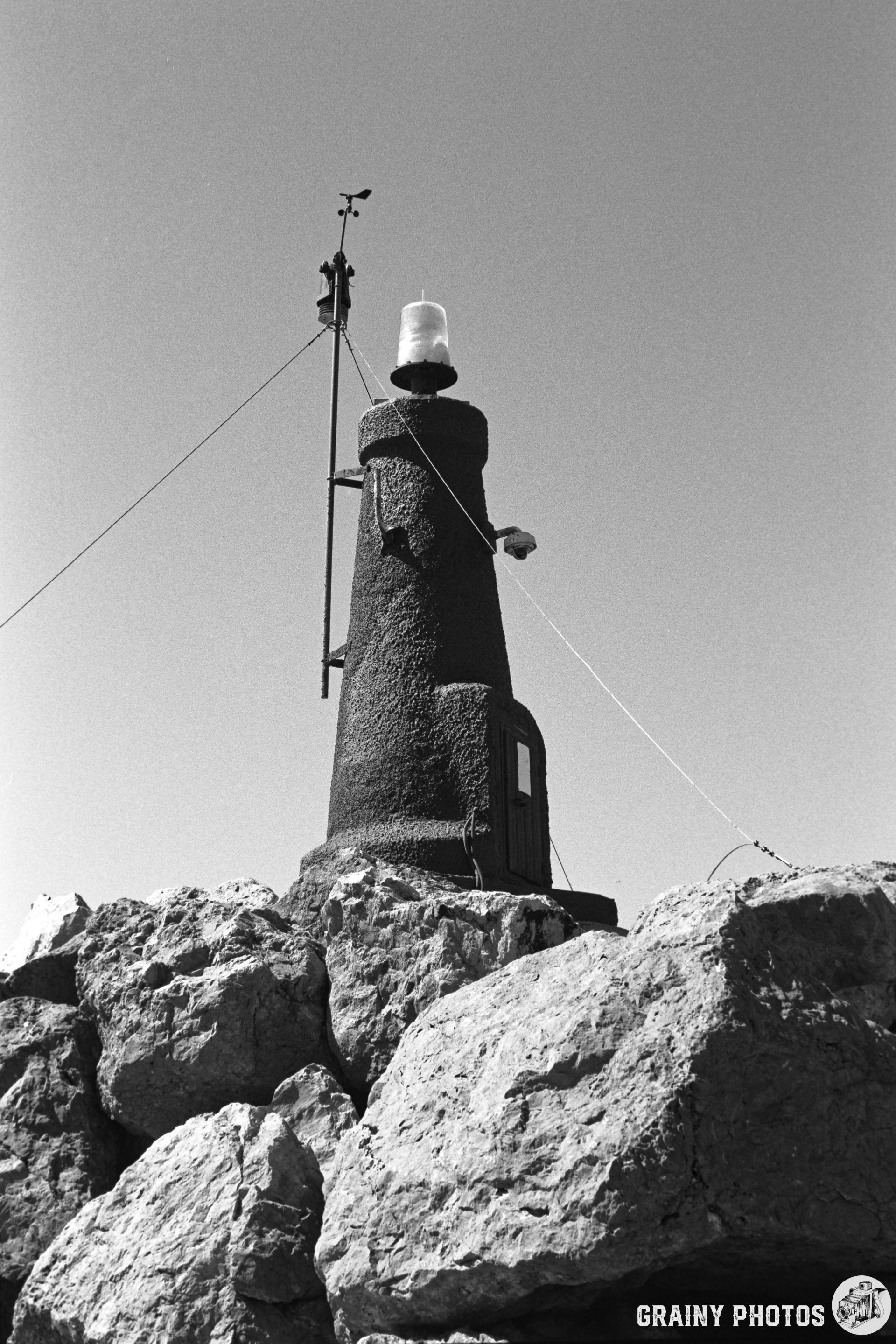 A black and white image of a lighthouse perched on large rocks, featuring a light beacon on top and a weather vane. The structure stands against a clear sky, emphasizing its significance as a navigational aid.