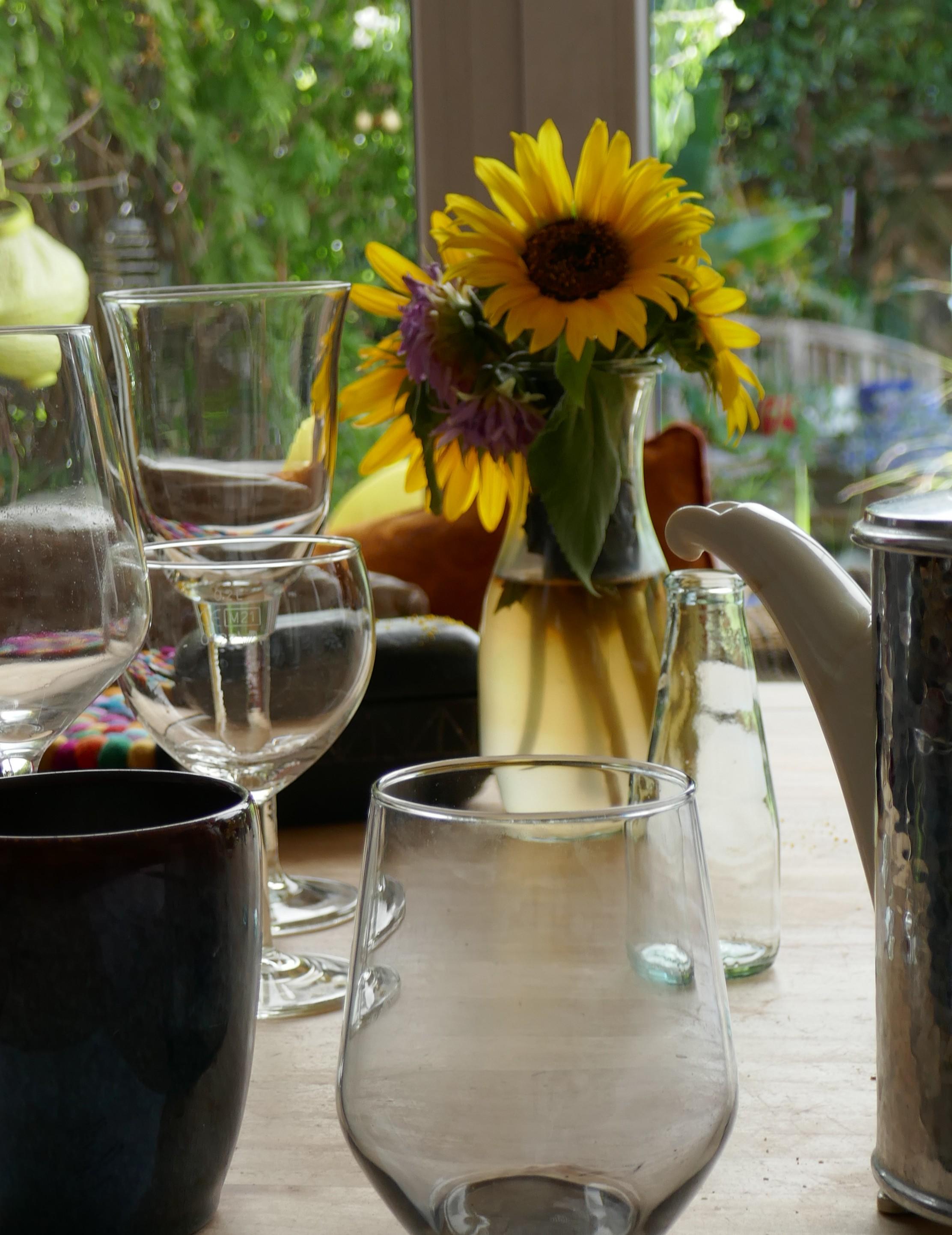 view over a table top, with a collection of empty clean wine glasses on the left and in the foreground, and a small vase with palmsized sunflowers arranged in it. there is also a lilac blossom. in the background a window frame, and the green of my garden forming a blurred wall of leaves. The water in the vase has a brown tint, showing a morbid accent as a contrast to the clear glass