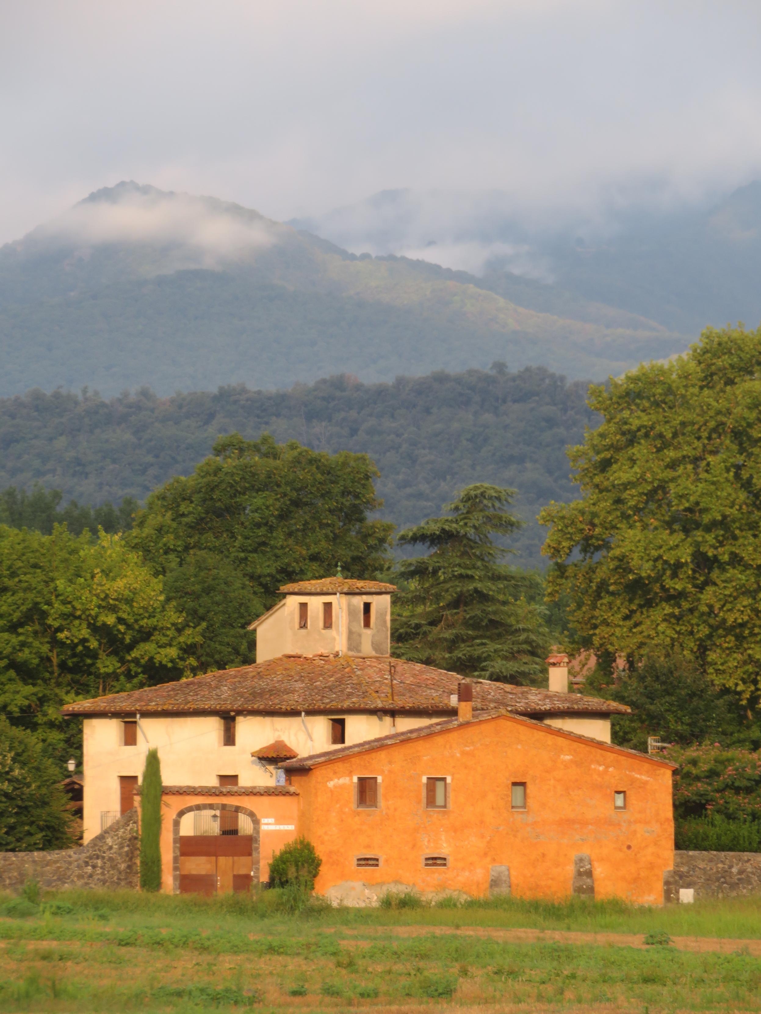 In the photograph taken today at dawn, a large "Masía", an old Catalan rural house, painted with light and warm colors, contrasts with the intense green of the surrounding forest and the blue-green of the mountain in the background, whose upper part is covered with clouds.