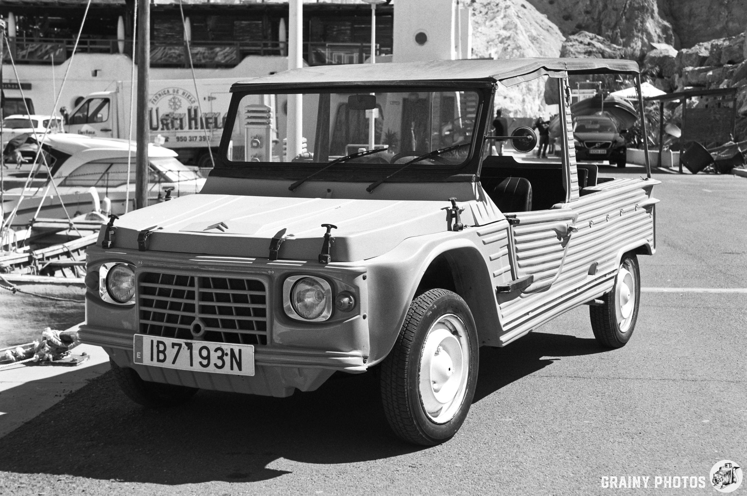 A classic light-colored utility vehicle parked near a marina, surrounded by boats, captured in black and white.