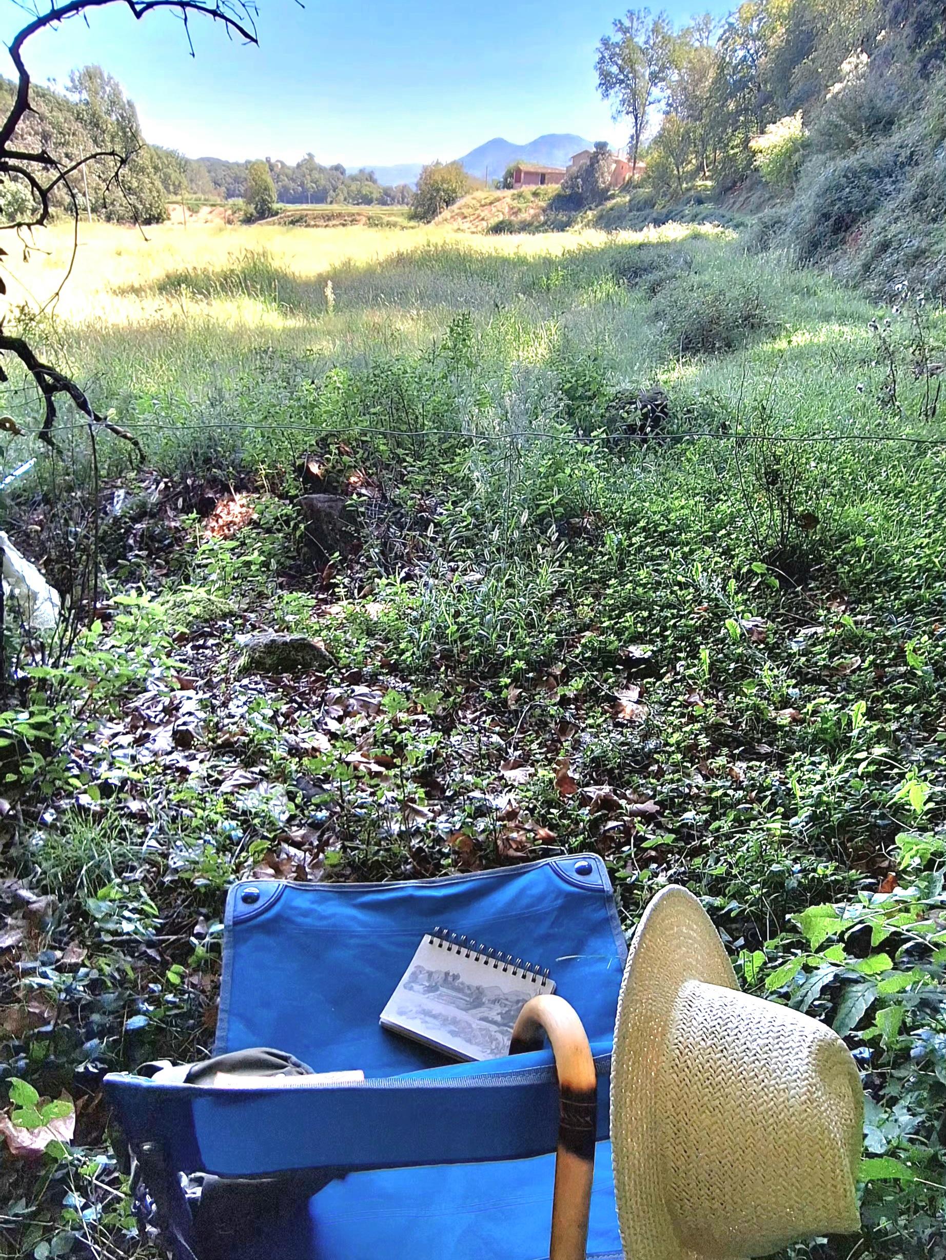 A folding chair, a walking stick, a hat, a bag with art supplies, and a sketchbook, sitting in the shade facing a beautiful landscape. The pleasure of drawing.