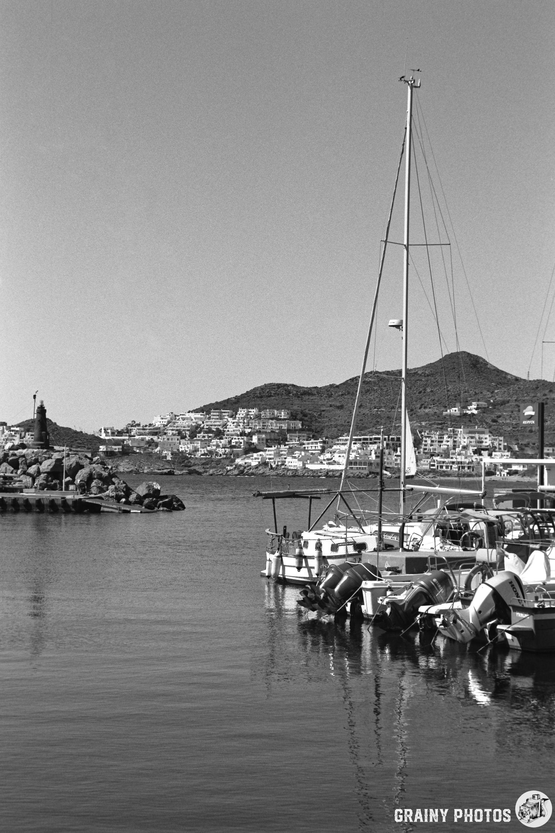 A serene black and white coastal scene featuring anchored boats in San José Marina, with a backdrop of hills and a small town. The calm water reflects the boats and the shoreline, creating a peaceful maritime atmosphere.