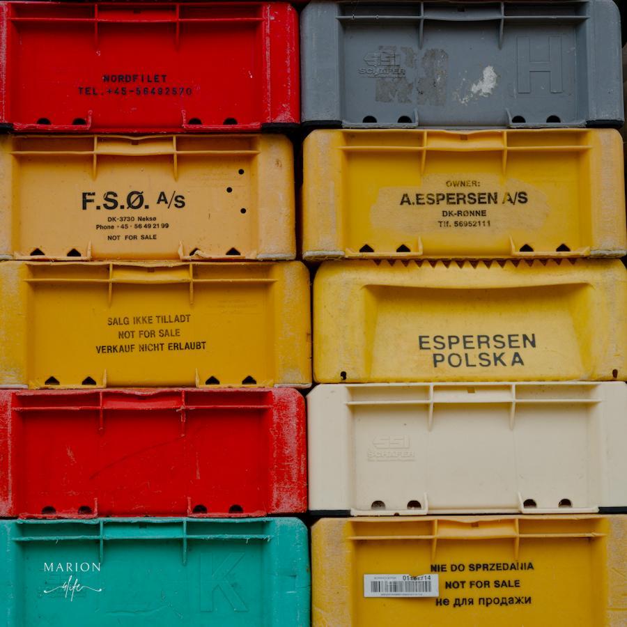 Es sind zwei Reihen mit je 5 unterschiedlich farbige Plastikkisten für den Fischtransport zu sehen, sie ergeben ein Quadrat. Auf den Kisten sind die jeweiligen Orte aufgedruckt, z. B. ist auf einer hellgelben Kiste F.S.O zu lesen oder auf einer anderen hellgelben Kiste "Espersen Polska". Auf der Kiste oben links, rot, steht "Nordfilet" und eine Telefonnummer