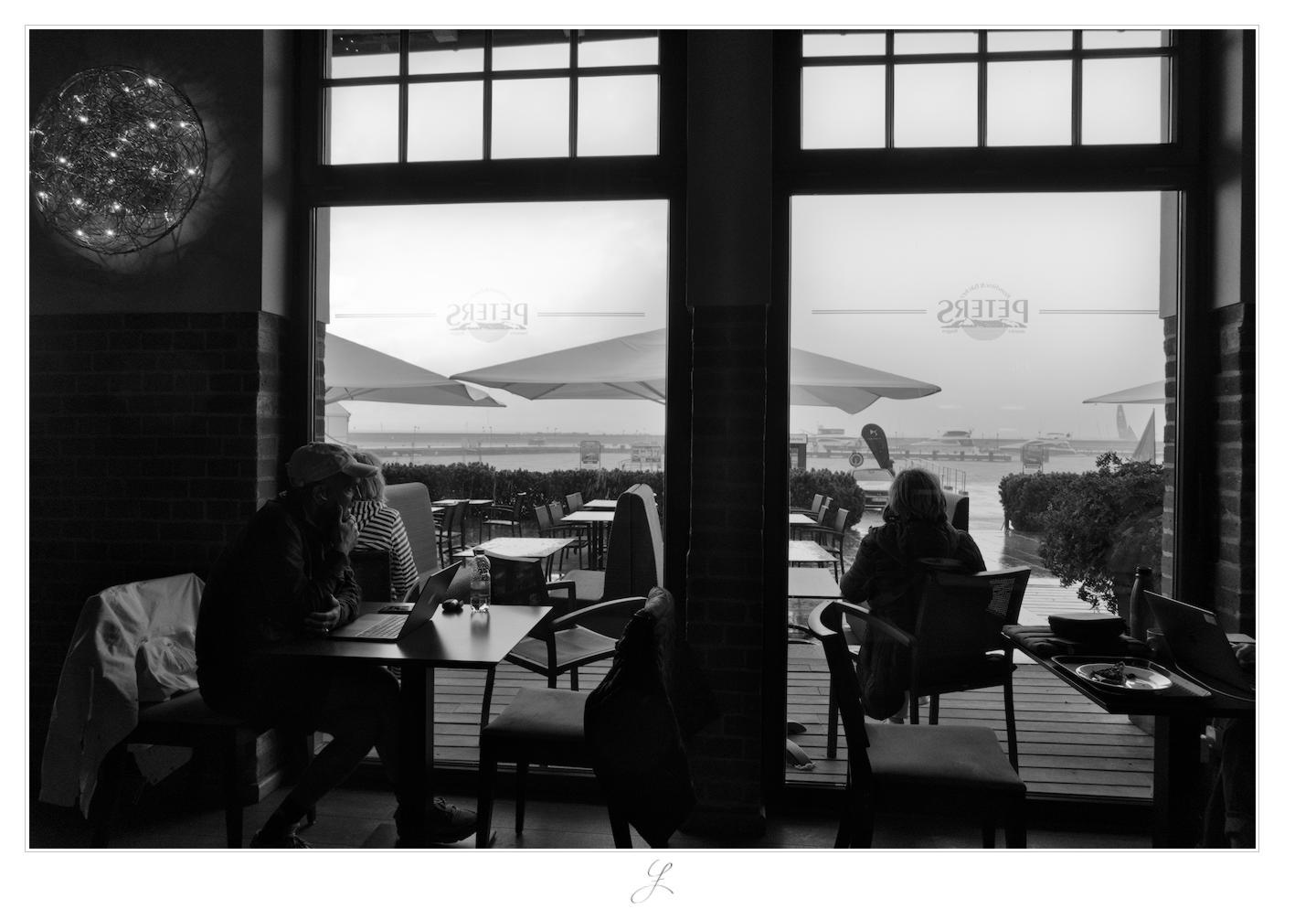 Dry Inside
From inside a café, the view extends outward through two large windows that reach down to the floor. Above, there are smaller windows with square-paned frames. To the left of the windows, a lamp with many small bulbs is mounted on the wall. The lower two-thirds of the wall are made of brick, the upper part is plastered. In front of the window and outside, there are many small tables with chairs. A man wearing a cap is sitting at a table in front of the left window, his laptop is on the tabletop. He is looking out at the heavy rain, his chin resting on his right hand. Outside, there are large parasols that eventually turned into large umbrellas.

AI disclaimer: Using my work, its meta data, written or derived description to create media with or train AI based systems is prohibited.