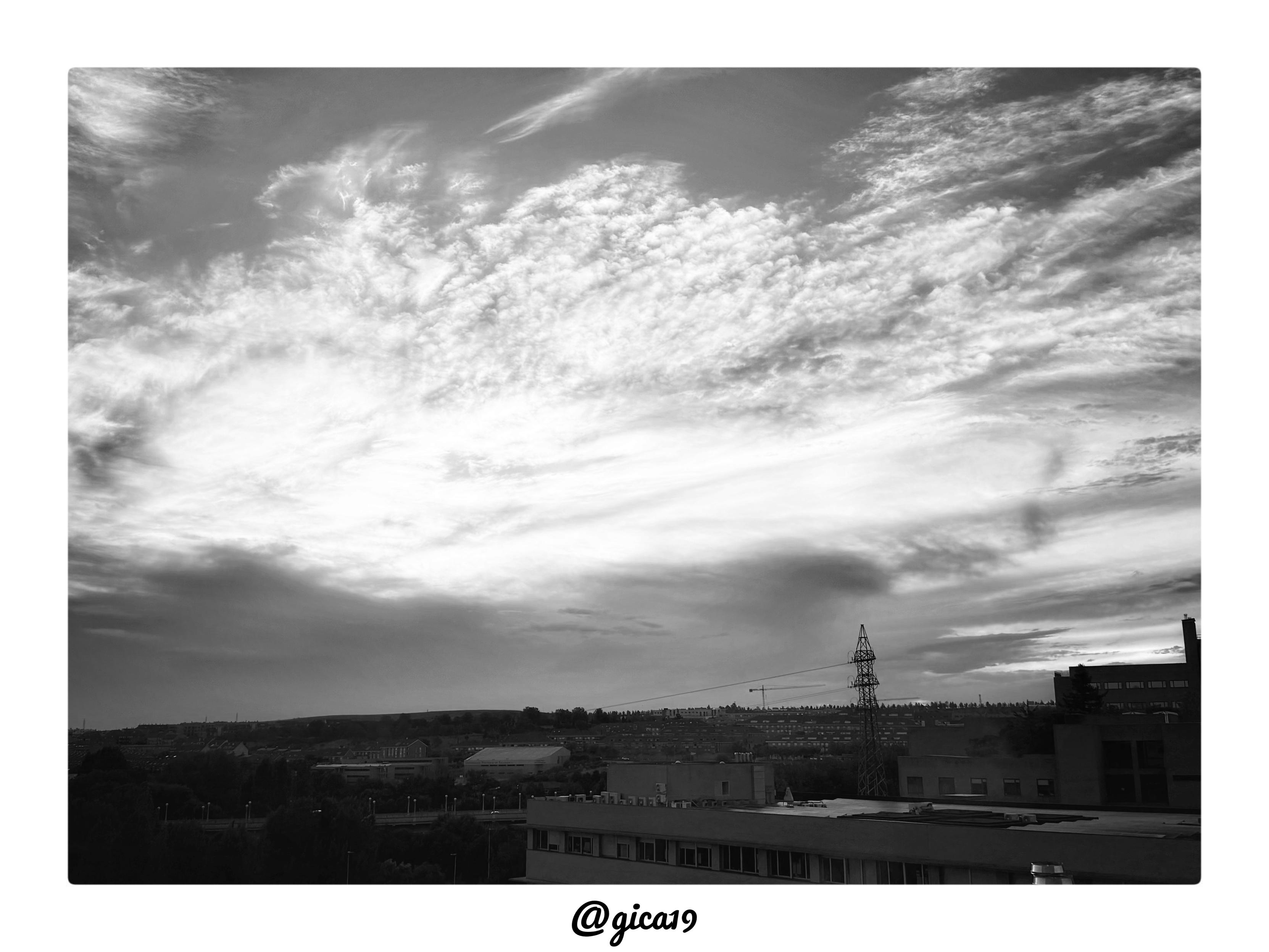 La imagen es una fotografía en blanco y negro de un cielo al atardecer, tomada desde la cuarta planta del Hospital Universitario de Salamanca.

El elemento protagonista es una extensa formación de nubes que ocupa aproximadamente dos tercios superiores del encuadre. Las nubes tienen una textura fibrosa y estriada, típica de nubes altas (cirros o altocúmulos), con un patrón que se despliega en diagonal desde la esquina superior izquierda hacia la derecha. La zona central de la masa nubosa está fuertemente iluminada, casi blanca pura, sugiriendo que el sol se encuentra oculto tras las nubes o muy cerca del horizonte, mientras que los bordes y las zonas periféricas muestran tonos grises más oscuros que dan volumen y profundidad dramática a la escena.

En la franja inferior de la imagen, mucho más oscura y en penumbra, se distingue:

- Una silueta de colinas u ondulaciones del terreno en el horizonte
- Una zona urbanizada con edificaciones residenciales de baja altura
- Un edificio en prime