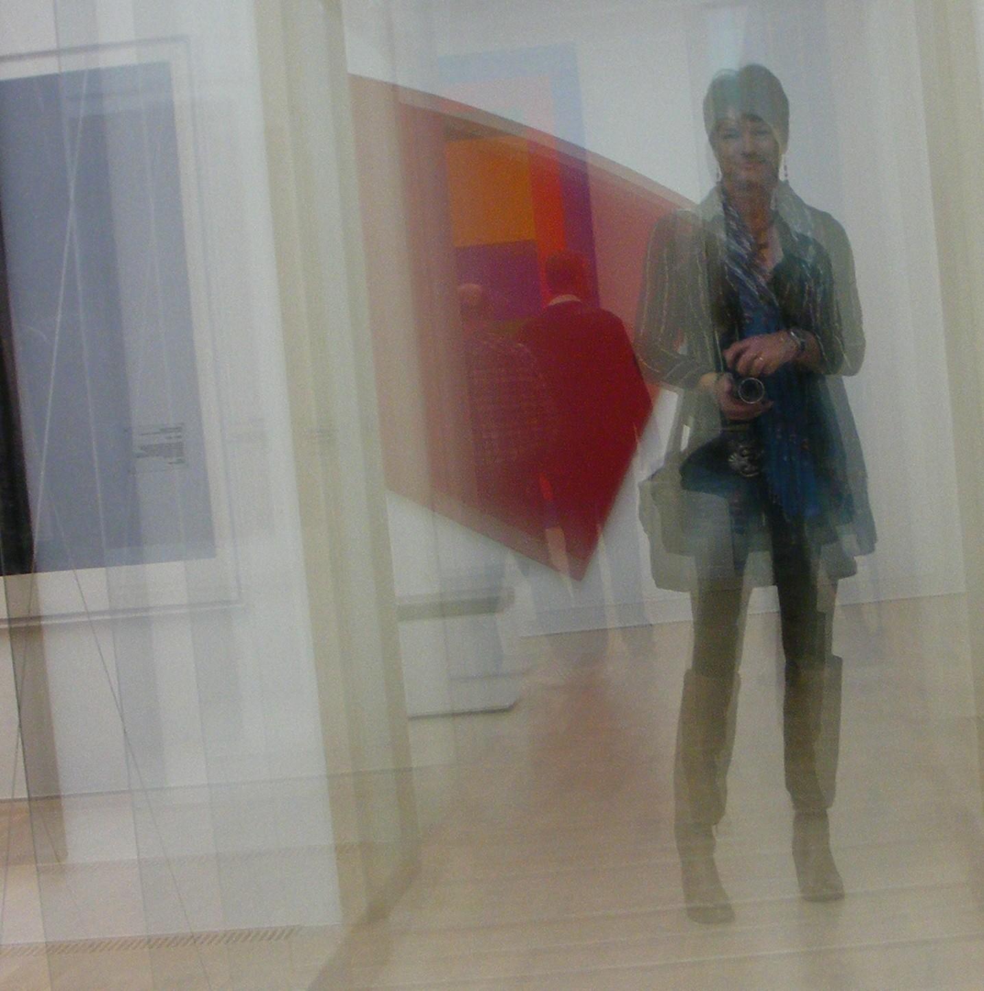 a square color photo showing blurred reflections of a fashionable woman with short hair, holding a camera at hip height, an angled corner of a wall, and a blurred transparent man walking towards a red swirl of color in the background.