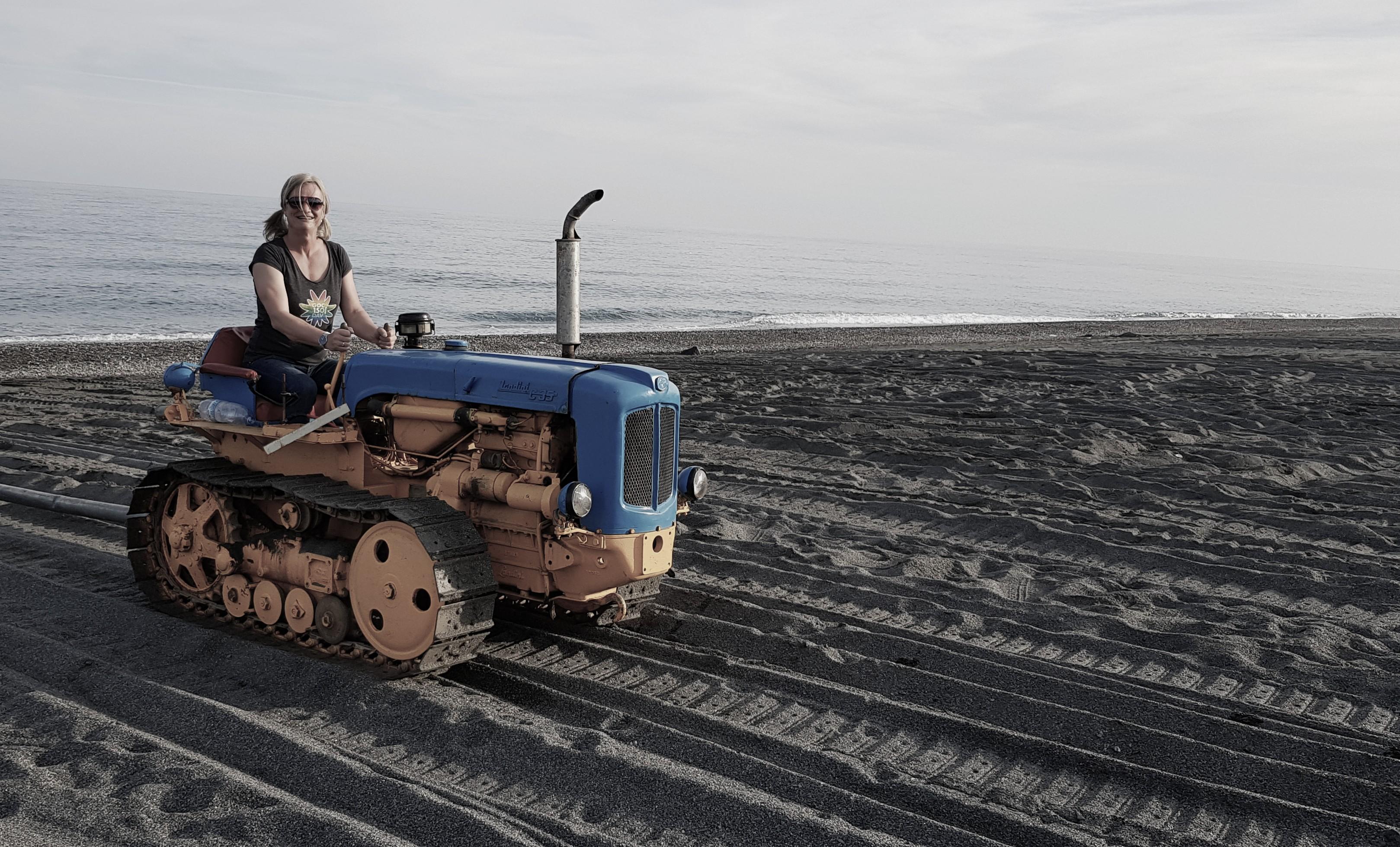 a caucasian woman sitting on a tracked tractor on a sandy beach. The tractor is quite small, just an angine with a hod, and a seat behind, plus the tracks on the side. It is yellow and blue. The sand is marked with multiple passes of the tractor having gone back and forth. The picture has faded colors, the left half shows the tractor, the right just beach, the sea, and the horizon blurring a faded blue sea into a faded sky.