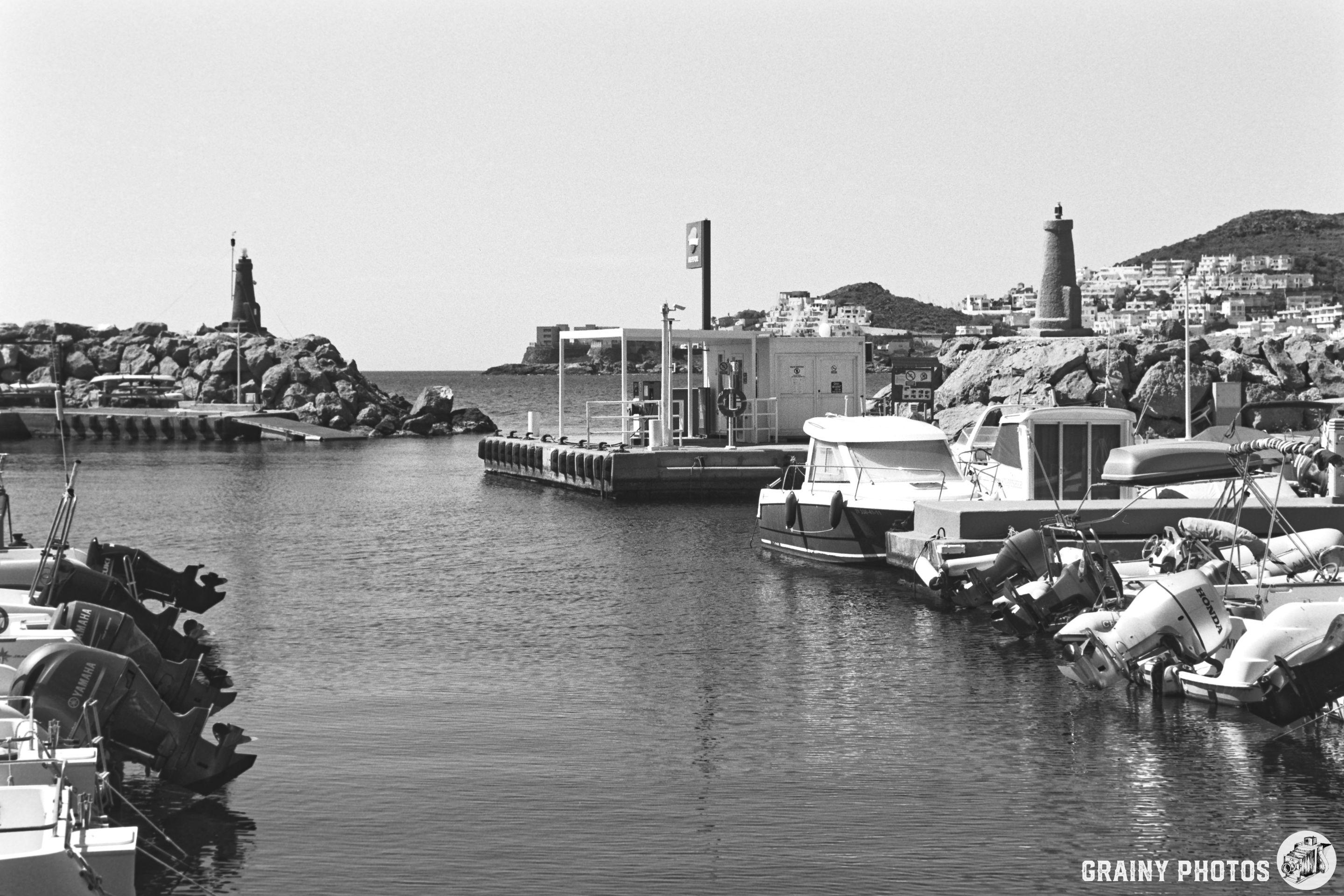 A monochrome image of San José Marina featuring numerous boats docked along the waterfront, with rocky shores and lighthouses visible in the background against a clear sky.