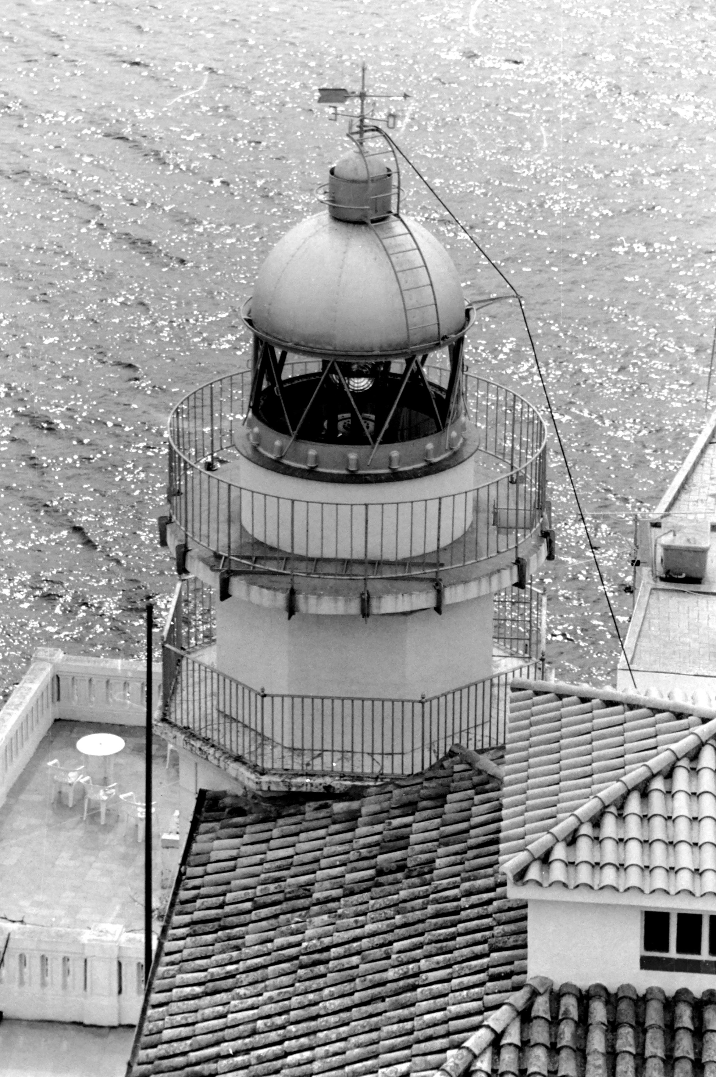 Black and white photograph, taken on film, of the Peñíscola Lighthouse from an "aerial" view from the terrace of the historic Peniscola Castle.
Built in the 19th century, its octagonal tower appears surrounded by the rooftops and terraces of the adjacent buildings. The shimmering Mediterranean Sea forms the background.