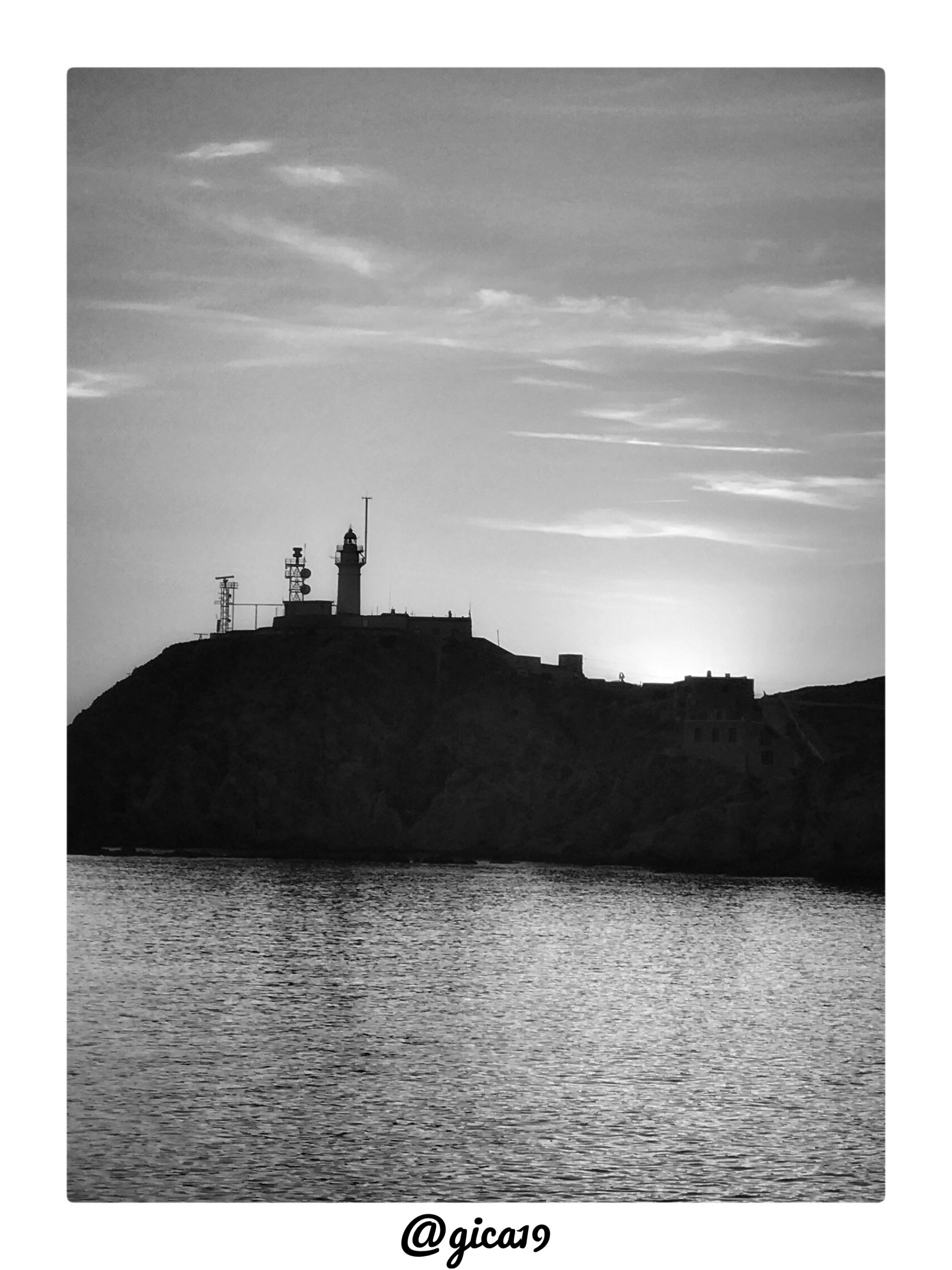 La fotografía, en blanco y negro, capta el Faro del Cabo de Gata recortado a contraluz sobre un cielo de atardecer, con el sol ocultándose justo detrás del promontorio y generando un halo de luz difusa que ilumina el horizonte desde el lado derecho de la imagen, señal del descenso solar hacia el mar.

El acantilado rocoso ocupa la franja central de la imagen y se presenta como una silueta oscura y compacta, con un perfil abrupto que cae directamente sobre el mar. Sobre la cima se distingue con claridad la torre del faro, de forma cilíndrica y esbelta, acompañada de varias antenas y estructuras auxiliares —entre ellas lo que parece un radar o equipo de comunicaciones— típicas de una instalación de señalización marítima activa. A la derecha del faro se aprecian construcciones anexas de perfil más bajo, posiblemente edificaciones del antiguo recinto o dependencias del propio faro.

La parte superior de la imagen está dominada por un cielo surcado por finas nubes alargadas dispuestas en ba