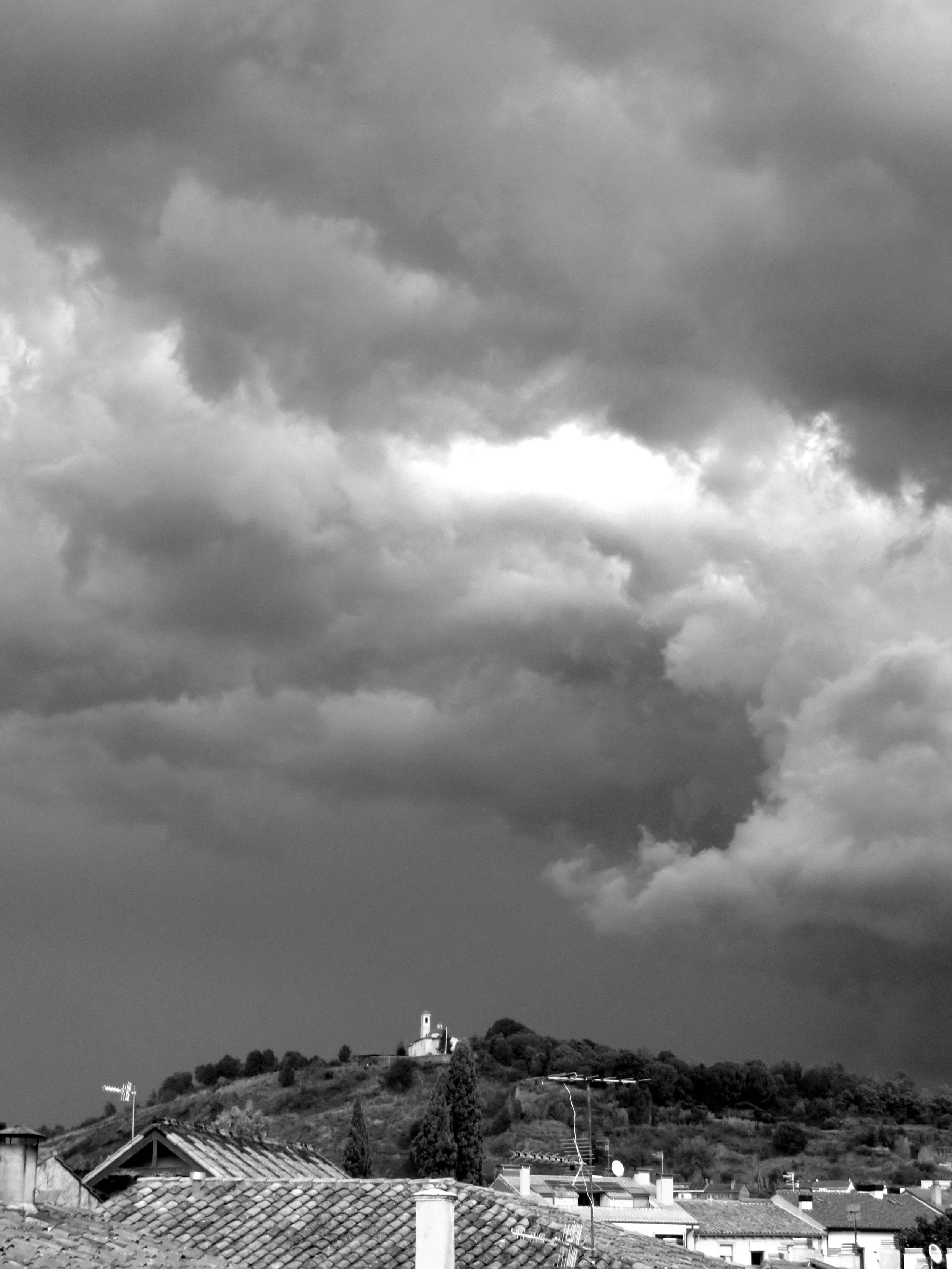 Black and white photograph of a stormy sky over the city of Olot, with the volcano and the hermitage of San Francesc in the background.
