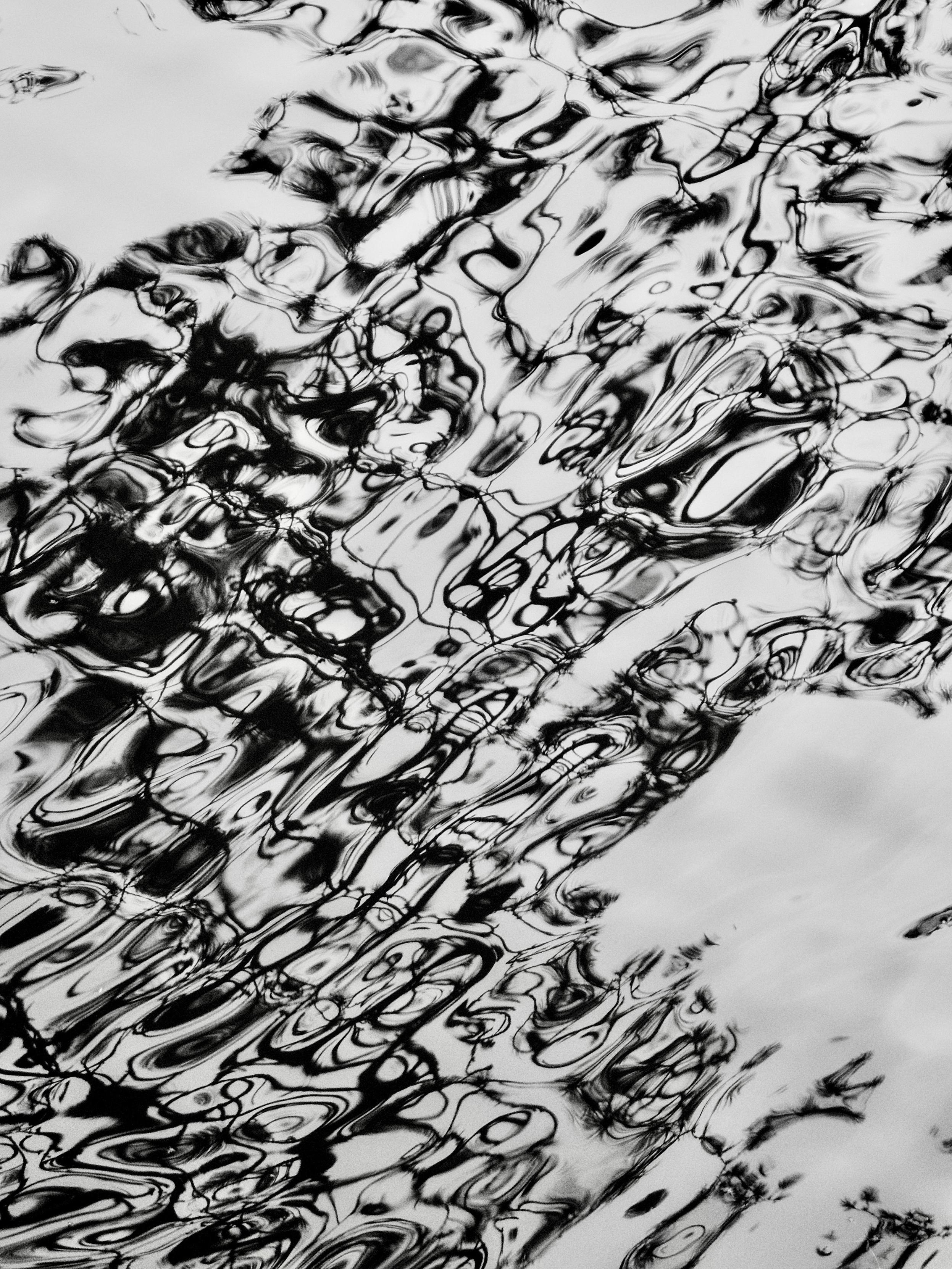 Abstract black and white image taken with an OM System Tough TG-7 pocket camera and showing the dark reflections of tree branches reflecting on the rippled surface of a large pond, where the movement of the wind-blown water has created abstract, dancing patterns out of the reflections.