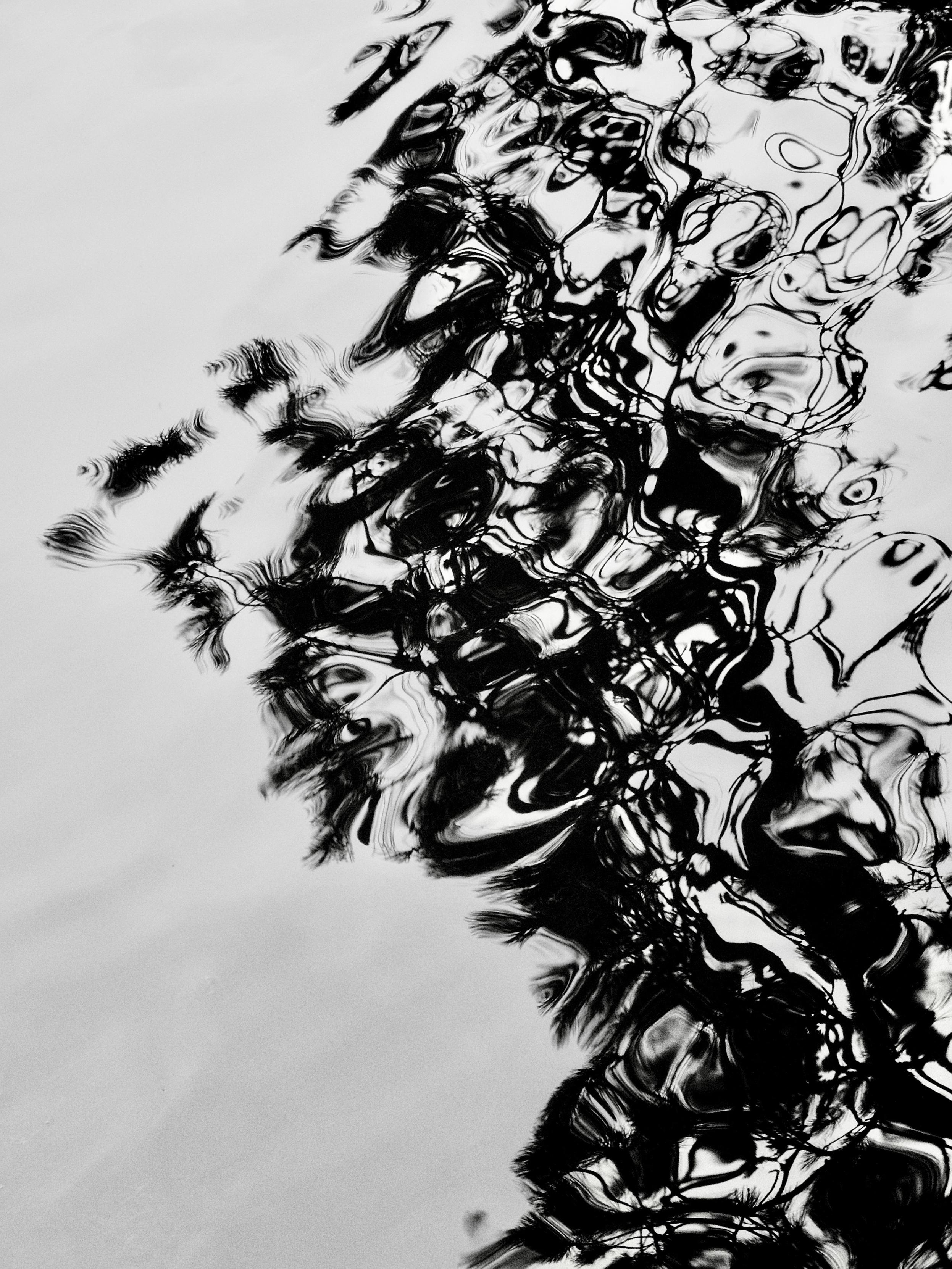 Abstract black and white image taken with an OM System Tough TG-7 pocket camera and showing the dark reflections of tree branches reflecting on the rippled surface of a large pond, where the movement of the wind-blown water has created abstract, dancing patterns out of the reflections.