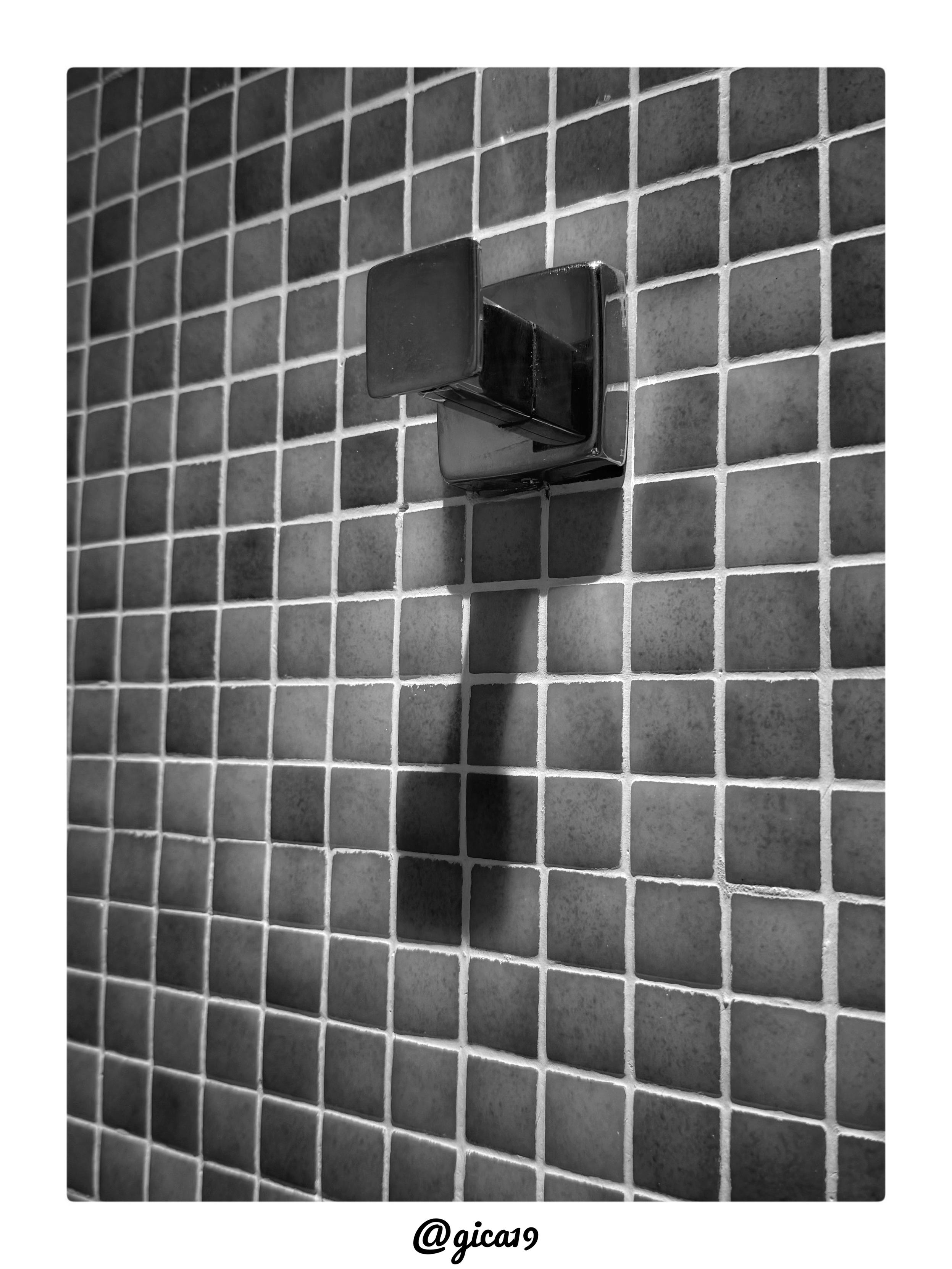 La imagen es una fotografía en blanco y negro, tomada en un ángulo ligeramente inclinado hacia arriba, que muestra una pared de baño o ducha completamente recubierta de azulejos pequeños y cuadrados de tamaño uniforme (tipo mosaico), separados por líneas de lechada blancas que forman una cuadrícula muy regular.

En el centro-superior de la composición hay un soporte para toallas o ropa, de metal cromado o acero pulido, con un diseño angular y geométrico compuesto por superficies planas que se pliegan formando un gancho o brazo abatible. Este tipo de accesorio suele estar articulado, permitiendo plegarlo contra la pared cuando no se usa y desplegarlo para colgar una toalla o prenda mientras se está en la ducha.

La luz incide de forma lateral, generando un reflejo intenso en las caras metálicas del soporte y proyectando una sombra alargada y bien definida hacia abajo sobre los azulejos, lo que acentúa el contraste característico del blanco y negro. La repetición geométrica de los azulej
