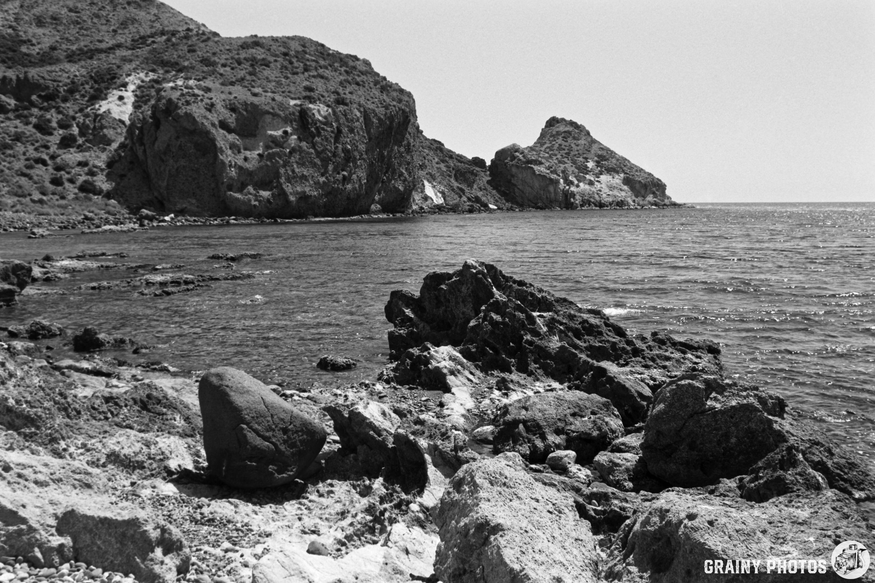 A serene black-and-white coastal landscape featuring rugged rocks along the shore, gentle waves lapping against the shoreline, and distant cliffs rising from the sea under a clear sky.