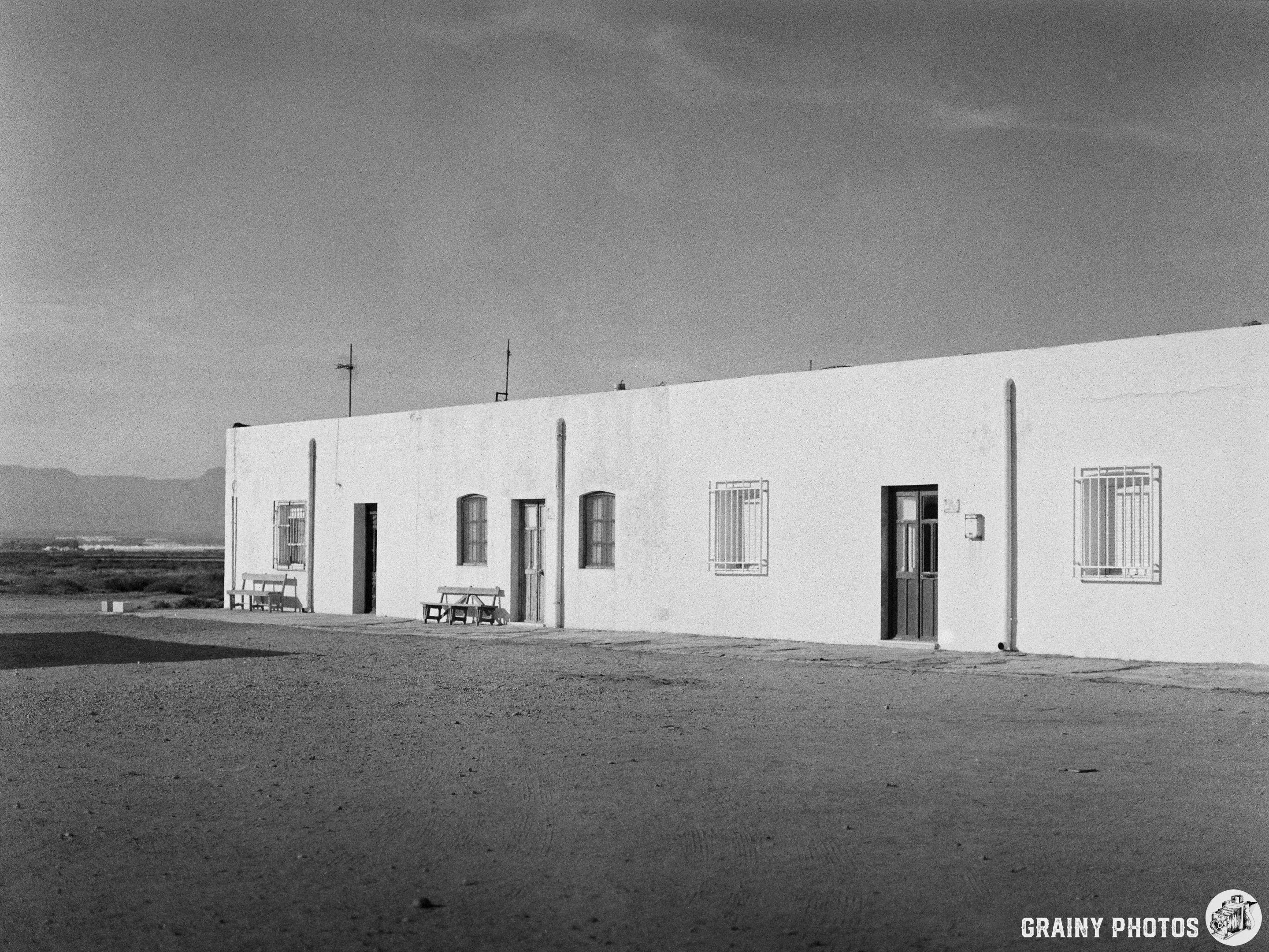 A monochrome image of a simple, low-rise Workers' cottages at Las Salinas. The structure is surrounded by a gravel area under a clear sky, suggesting a remote setting. Two wooden tables and chairs are visible outside the building.