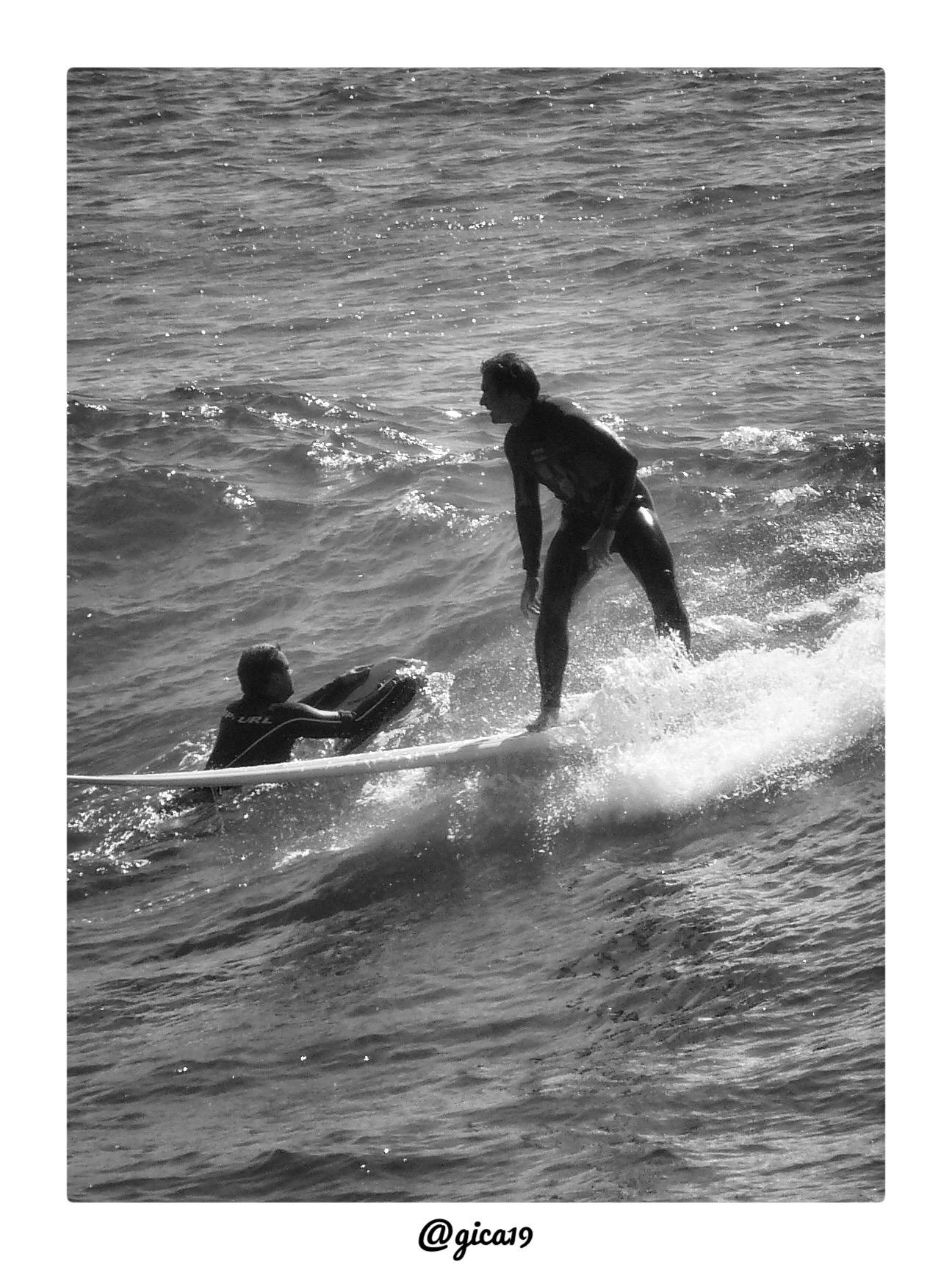 La imagen, tomada en las playas de Aveiro, Portugal, es una fotografía en blanco y negro que capta un momento de surf.

En primer plano y algo a la izquierda, un surfista está tumbado boca abajo sobre su tabla, remando o esperando en el agua, con el brazo extendido hacia adelante. Detrás de él, y ocupando el centro de la imagen, otro surfista se encuentra de pie sobre su tabla, en plena maniobra de deslizamiento, con las rodillas ligeramente flexionadas y los brazos abiertos para mantener el equilibrio mientras cabalga una ola pequeña que rompe formando espuma blanca.

El mar presenta un oleaje suave con la superficie rizada, típica de las condiciones atlánticas de la costa portuguesa. El contraste en blanco y negro resalta el brillo del agua salpicado de destellos de luz, sugiriendo una hora del día con sol intenso, probablemente cerca del mediodía o primeras horas de la tarde. La espuma de la ola rompiente destaca en tonos muy claros frente al gris más oscuro del mar de fondo, aporta