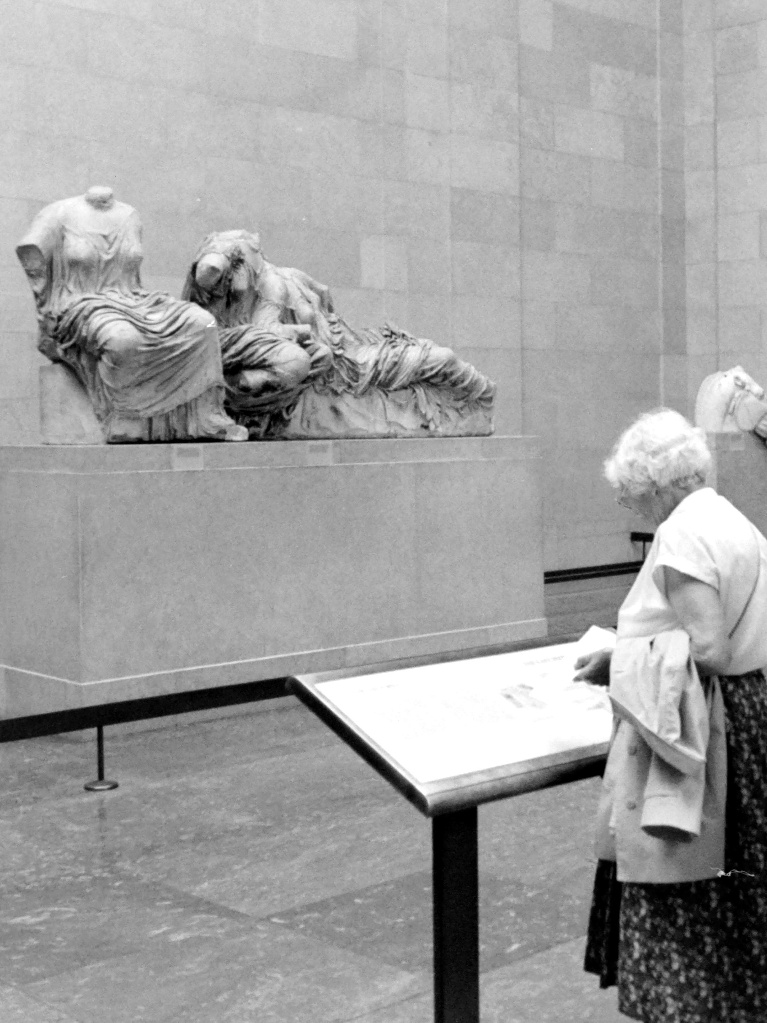 The "analog" black and white photograph in 3:4 portrait format shows a visitor in a room of the British Museum in front of The Three Goddesses, a group of marble statues from the East Pediment of the Parthenon in Athens, created by the sculptor Phidias between 438 and 432 BC. They are traditionally identified from left to right as Hestia (goddess of the hearth), Dione, and her daughter Aphrodite (goddess of love). An alternative theory suggests they represent Thalassa (the Sea) in the lap of Gaia (the Earth).