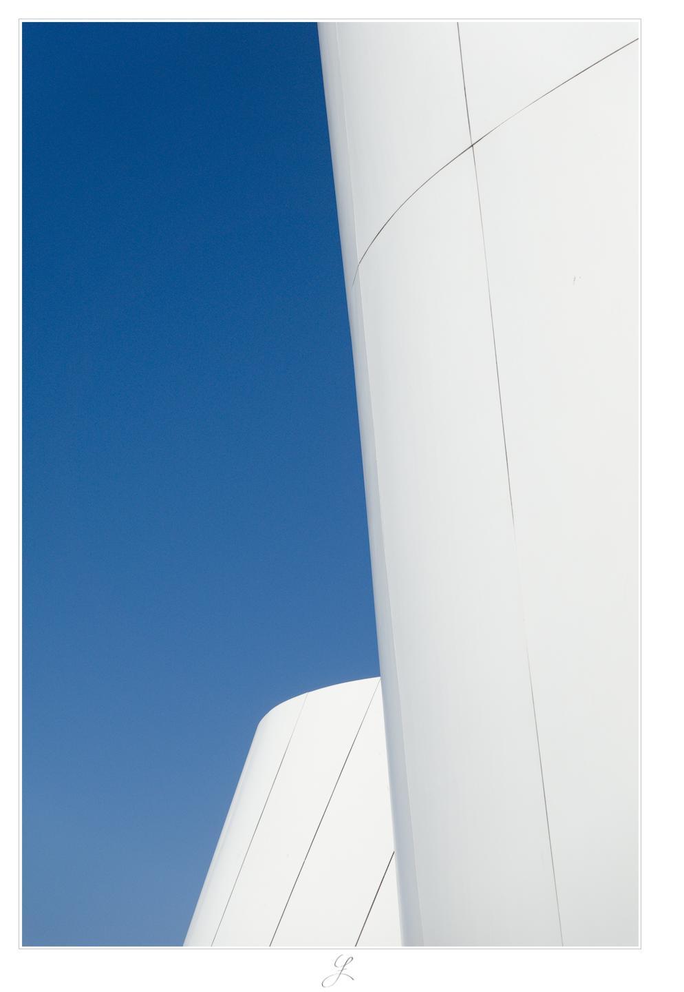 The portrait-oriented image shows two white facades against a cloudless, light-blue sky. Both facades consist of large, rectangular elements and have a curved, rounded outline. Thin black lines mark the joints between the facade elements. The facade of the building in the foreground leans to the left and fills roughly the right half of the scene diagonally. Behind it is the second, significantly lower building, whose facade leans to the right and disappears behind the building in the foreground. The image consists mainly of brilliant white and vivid blue.

AI disclaimer: Using my work, its meta data, written or derived description to create media with or train AI based systems is prohibited.