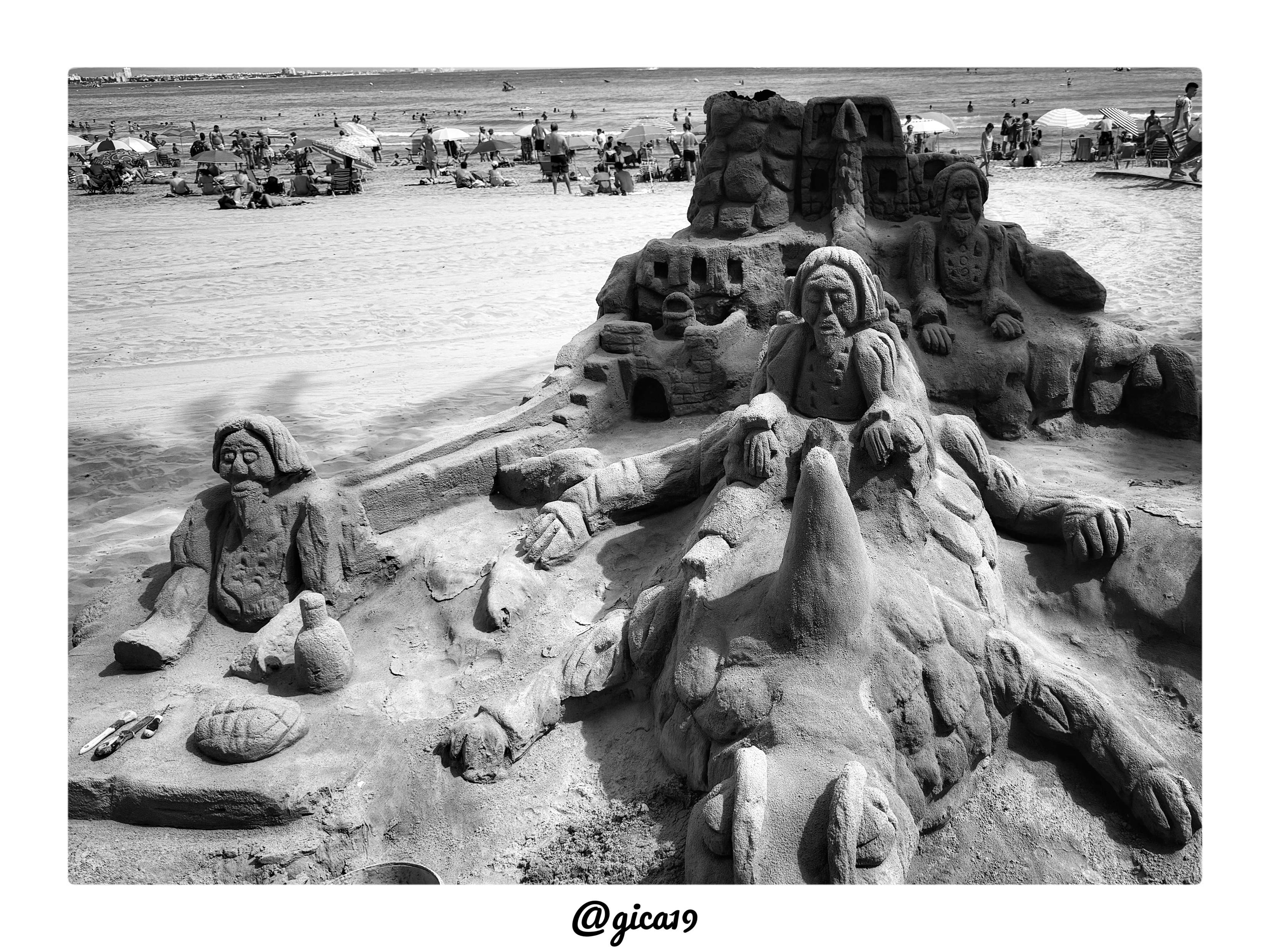 La imagen es una fotografía en blanco y negro tomada en una playa concurrida, mostrando un gran castillo de arena esculpido con figuras humanas detalladas.

La escena está tomada desde un ángulo bajo y elevado a la vez, que da protagonismo a una escultura de arena monumental en primer plano mientras la playa y el mar se extienden como telón de fondo. Al fondo se aprecia una línea de costa con edificios y montañas difuminadas por la calima, además de numerosas personas bañándose en el mar y sentadas bajo sombrillas en la arena.

El elemento central es una obra escultórica muy elaborada que representa varias figuras humanas con rasgos envejecidos: rostros barbudos, ojos cerrados o entornados, y ropajes con pliegues tallados con gran detalle, en un estilo que recuerda a personajes bíblicos o de una escena narrativa clásica. Detrás de las figuras se eleva una estructura que simula un castillo o fortificación, con torres, ventanas en arco y muros de piedra tallados en la arena, dando profun