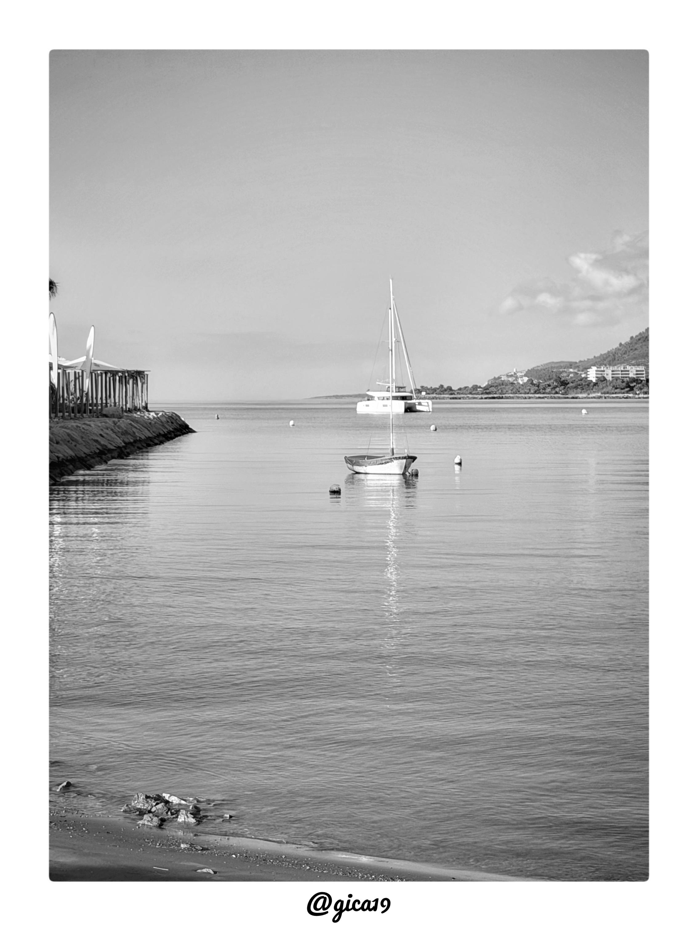 La imagen es una fotografía en blanco y negro tomada por la mañana en la Playa Sur de Peñíscola, con una composición serena y muy equilibrada centrada en el agua en calma.

En primer plano se extiende una playa de arena con algunas piedras dispersas cerca de la orilla, donde el agua llega con un movimiento suave, casi sin oleaje. El mar ocupa la mayor parte del encuadre, mostrando una superficie prácticamente en calma que refleja con nitidez la luz del cielo y las embarcaciones ancladas.
A la izquierda de la imagen se distingue un espigón o rompeolas de piedra que se adentra en el mar, junto al cual hay una estructura de madera —posiblemente un chiringuito o zona de descanso techada— y varias palmeras que enmarcan la escena por ese lado, aportando un toque característico del paisaje mediterráneo.

En el centro de la composición, casi como punto focal, aparecen dos embarcaciones fondeadas: un pequeño velero de casco oscuro en primer término y, ligeramente más atrás, un catamarán de mayo