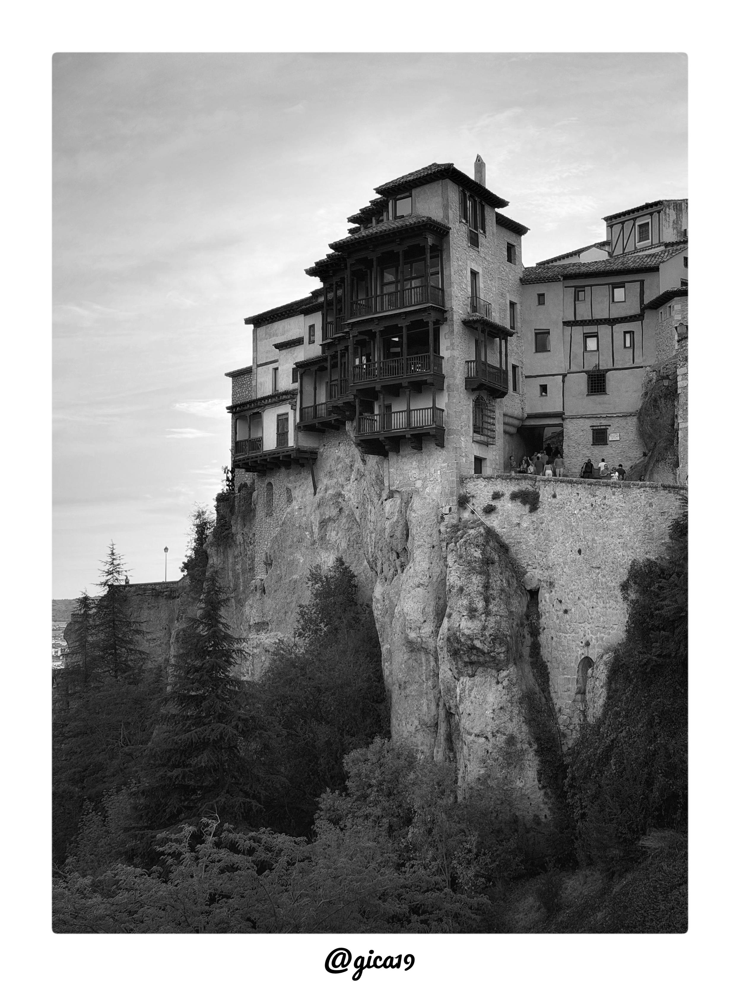 Esta es una fotografía en blanco y negro de las Casas Colgadas de Cuenca, uno de los símbolos más reconocibles de la ciudad y de la arquitectura tradicional española, capturada probablemente desde el mirador cercano al Puente de San Pablo.

La imagen se centra en el volumen principal de las Casas Colgadas, mostrando su característica estructura de balconadas de madera voladas que se asoman literalmente al vacío sobre la hoz del río Huécar. El encuadre vertical enfatiza la verticalidad de la roca caliza sobre la que se asienta el edificio, dejando un amplio espacio de cielo en la parte superior que aporta sensación de altura y aislamiento a la construcción.

Se distinguen con claridad los distintos cuerpos de balcones de madera oscura superpuestos en varios niveles, apoyados en ménsulas y vigas que sobresalen del muro de piedra y mampostería encalada. A la derecha se aprecia otra edificación con entramado de madera visible (estilo similar al de las casas tradicionales conquenses), y se 