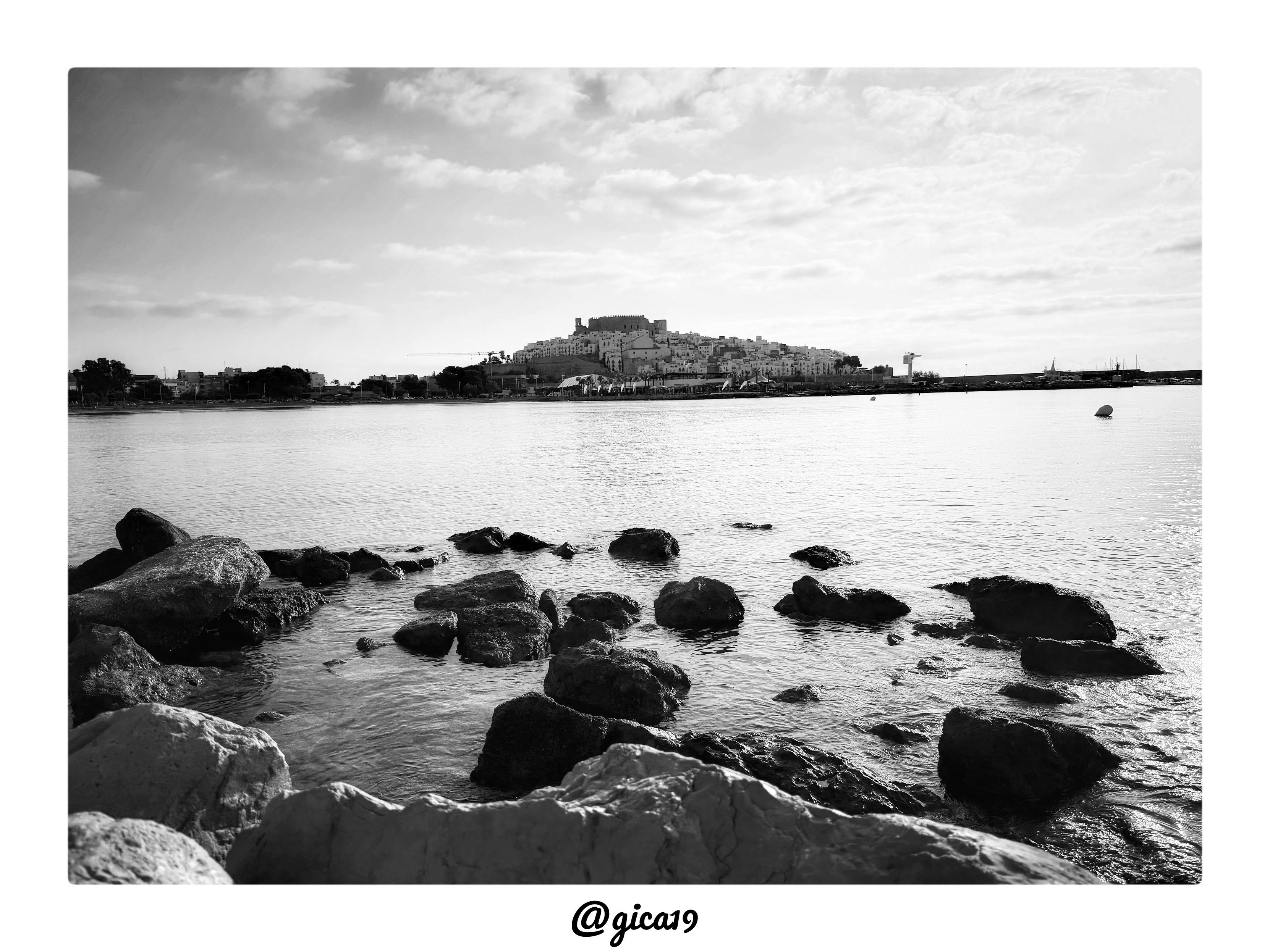 La fotografía es una composición marina en blanco y negro que muestra el Castillo del Papa Luna de Peñíscola visto desde la Playa Sur, poco después del amanecer.

La imagen sigue una estructura de tres planos clásica en fotografía de paisaje. En primer término, ocupando casi un tercio inferior del encuadre, aparece un grupo de rocas oscuras y de superficie irregular parcialmente sumergidas en el agua, que actúan como elemento de anclaje visual y aportan textura y profundidad. El plano medio lo ocupa una extensa superficie de agua en calma, con reflejos suaves de la luz matutina que crean pequeños destellos plateados sobre la superficie. Al fondo, en el tercio superior, se recorta la silueta del casco antiguo de Peñíscola sobre su promontorio rocoso, coronado por el castillo.

El Castillo del Papa Luna se distingue en lo alto del peñón, con sus torres y murallas almenadas dominando el perfil de la localidad. A su alrededor se despliega el conjunto de casas blancas encaladas del casco hi
